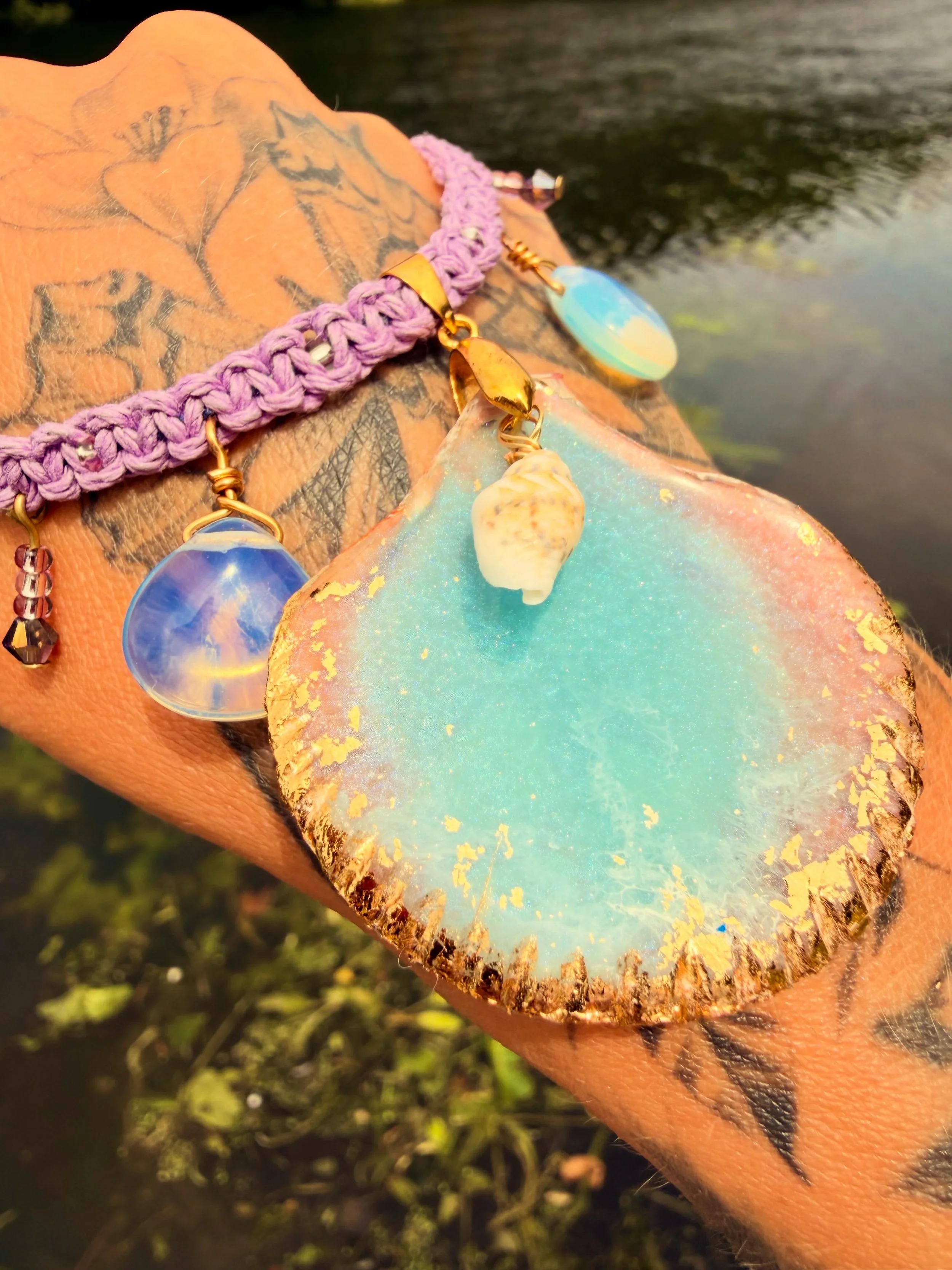 Close-up of a person's hand holding a large iridescent leaf with gold flecks, decorated with a lavender woven choker necklace with various colorful charms including a seashell and gemstone pendants.