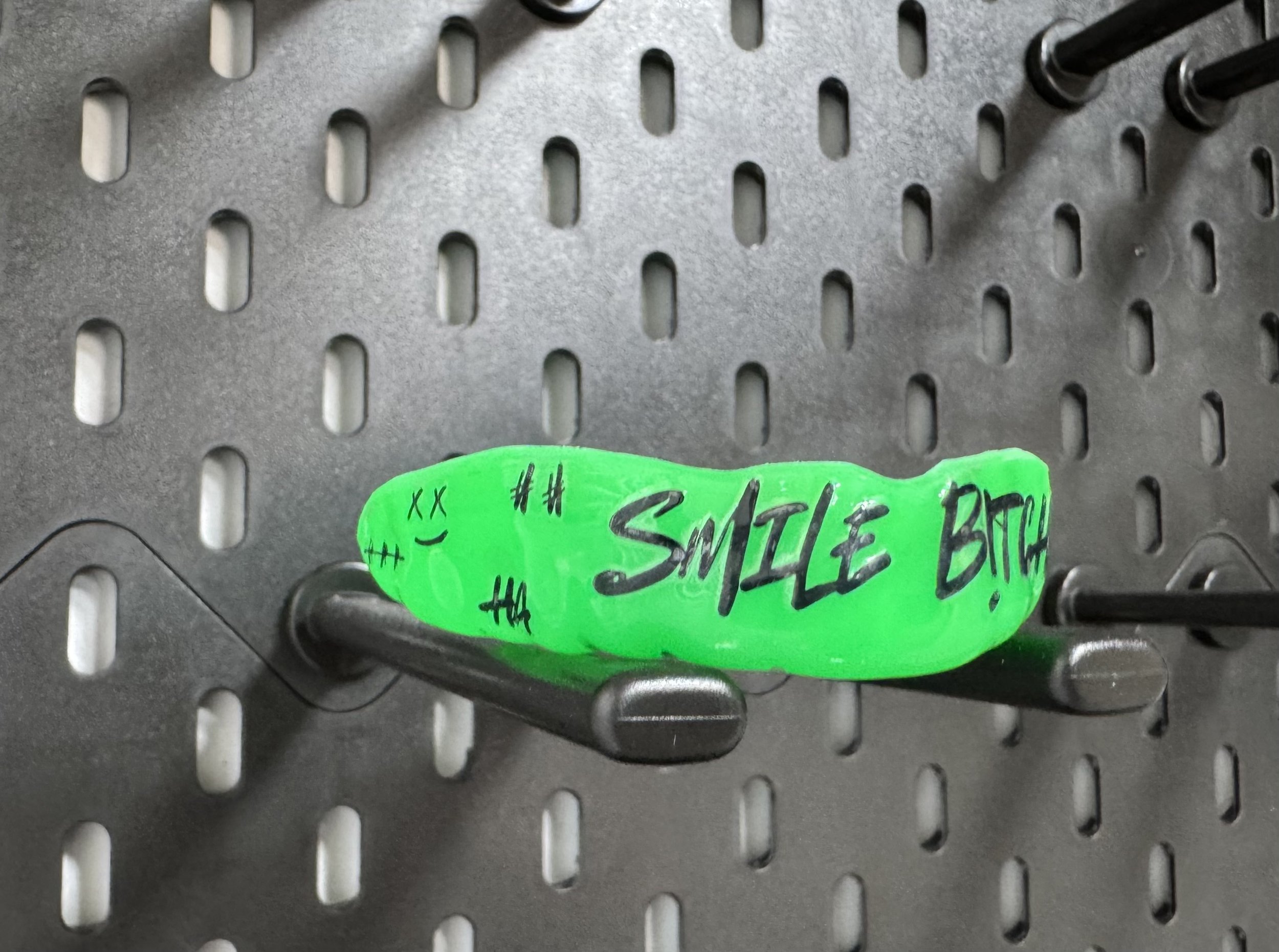 Groene Gebitsbeschermer met de woorden 'SMILE BITCH' en een lachend gezichtje, bevestigd aan een metalen houder tegen een geperforeerde metalen achtergrond.