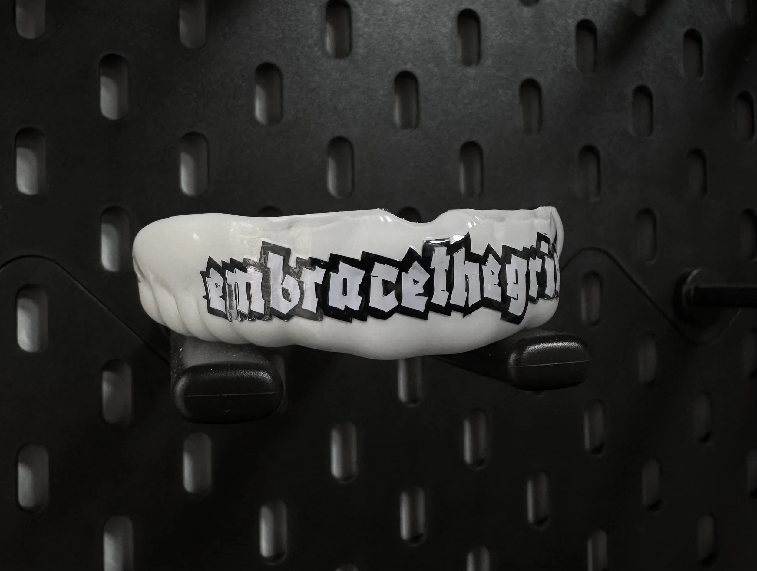 Een witte gebitsbeschermer met de zin 'embrace the grind' in zwarte, gotische letters, bevestigd aan een zwart geperforeerd vlak.