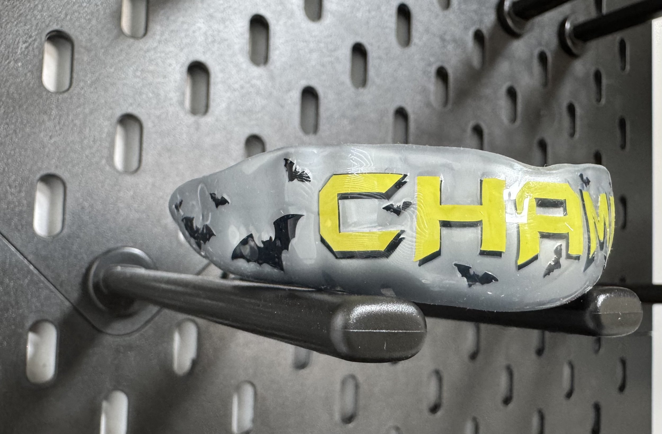 Een gebitsbeschermer met de tekst 'CHAMP' in gele en zwarte letters, gemonteerd op een metalen houder tegen een geperforeerde metalen achtergrond. En het design van Batman