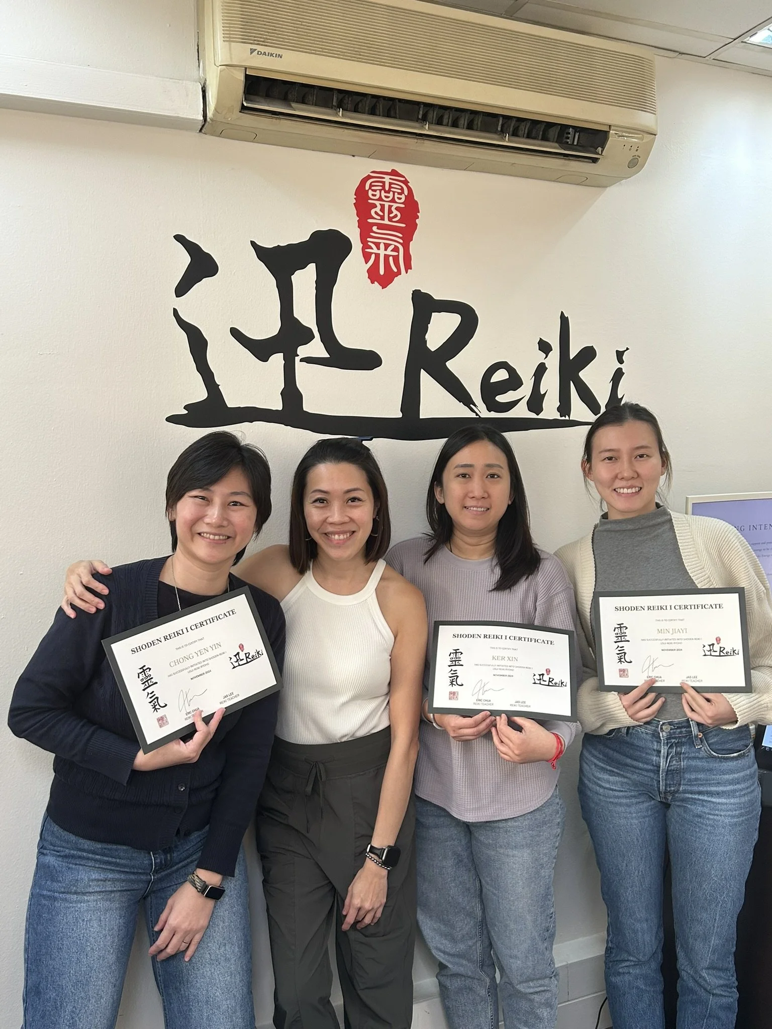 Four women standing in front of a wall with the words 'Reiki' and Japanese characters, holding certificates and smiling.