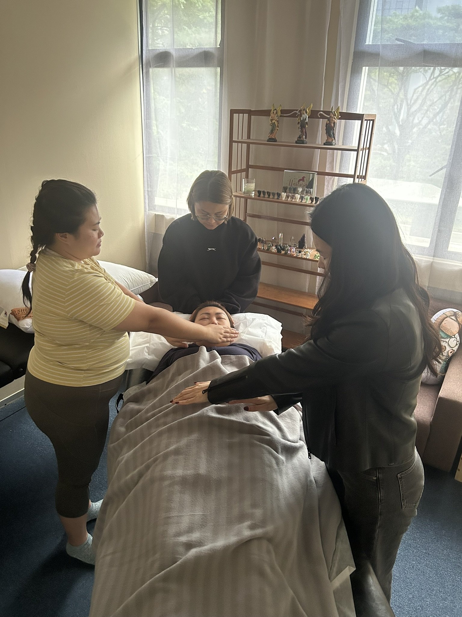 Four women in a room around a person lying in bed, with one woman touching the person's face, and another holding their hand, in a supportive setting.