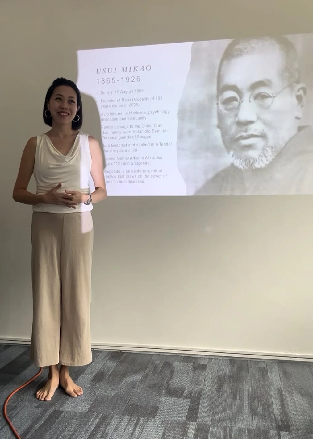 A woman standing barefoot in front of a projected presentation about Usui Mikao, with a smile, wearing a sleeveless cream-colored top and beige wide-leg pants, gesturing with her hands.