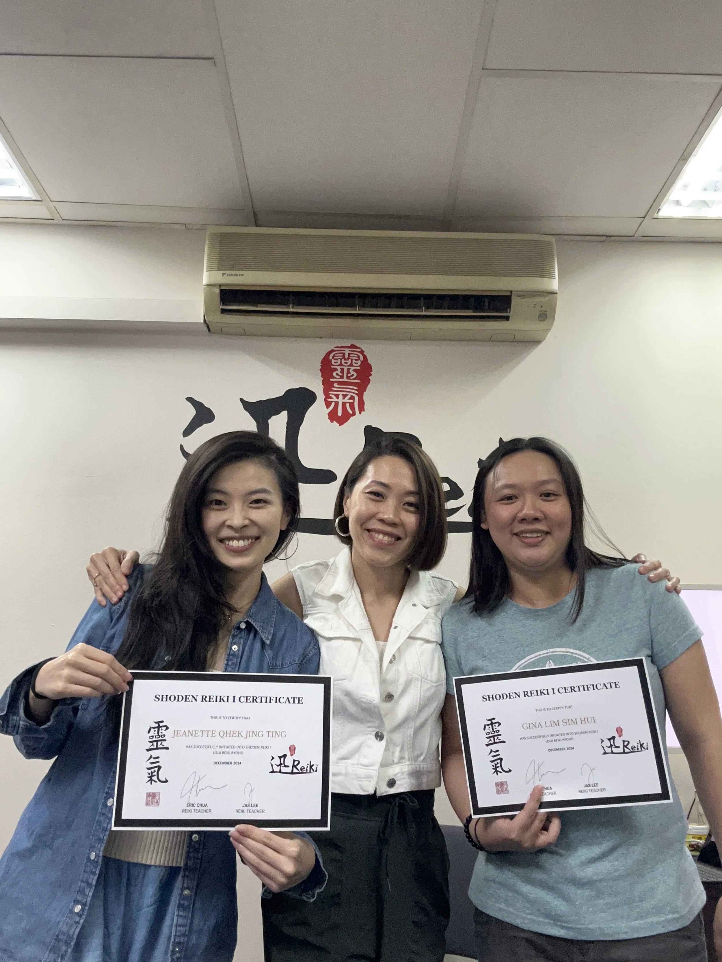 Three smiling women standing close together, two holding certificates, in an indoor setting with a white wall and Asian script behind them.