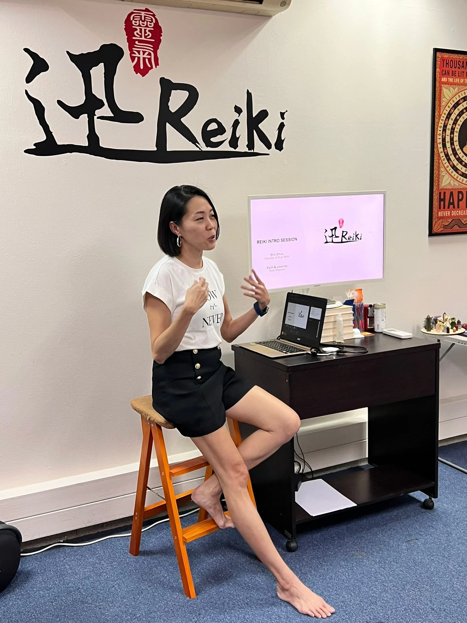 Woman giving a presentation in a room with a sign that says 'Reiki' on the wall and a computer on a desk.