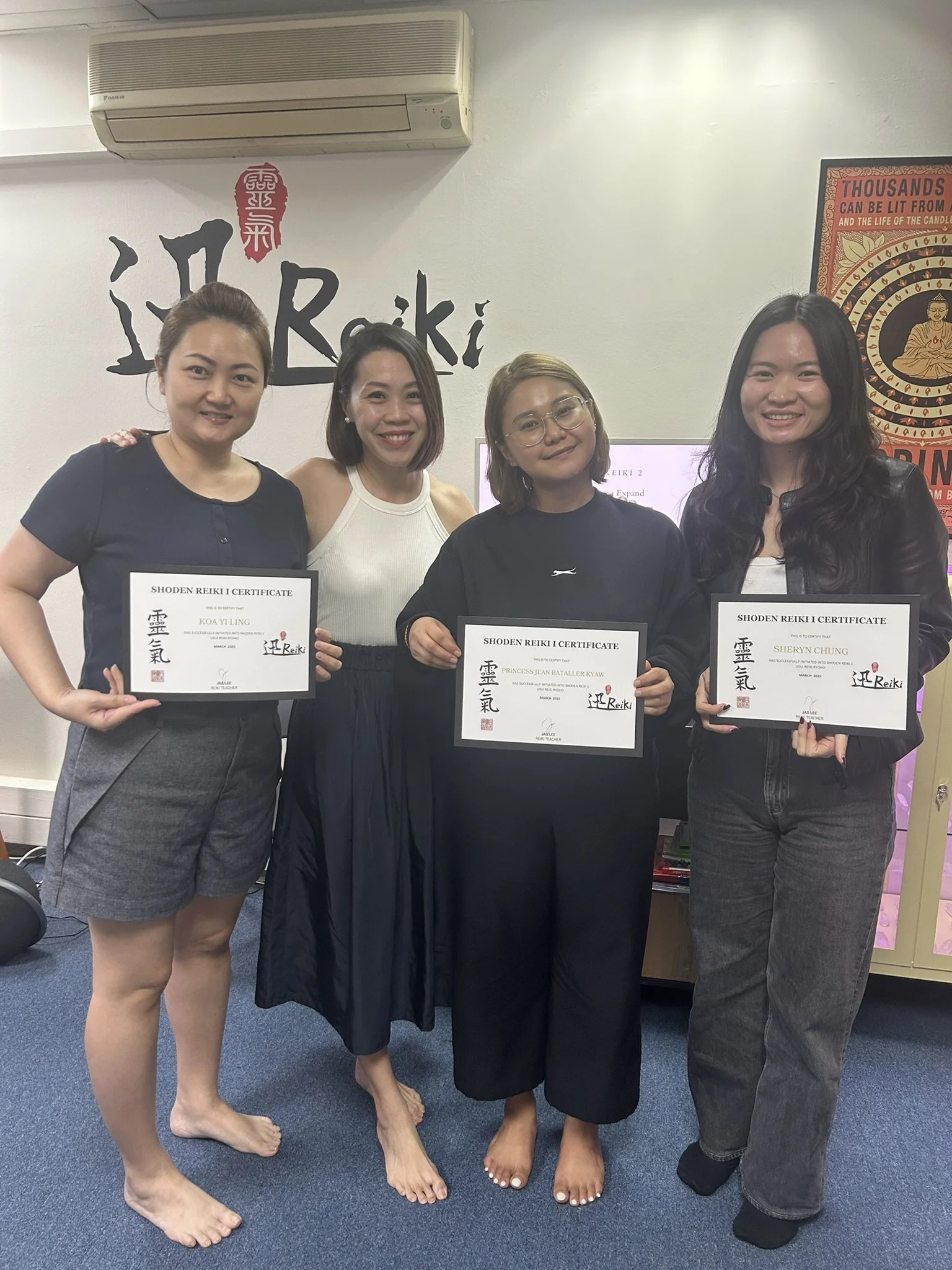 Four women standing together indoors, holding certificates, smiling, with a wall sign reading 'Reiki' behind them. All are barefoot.