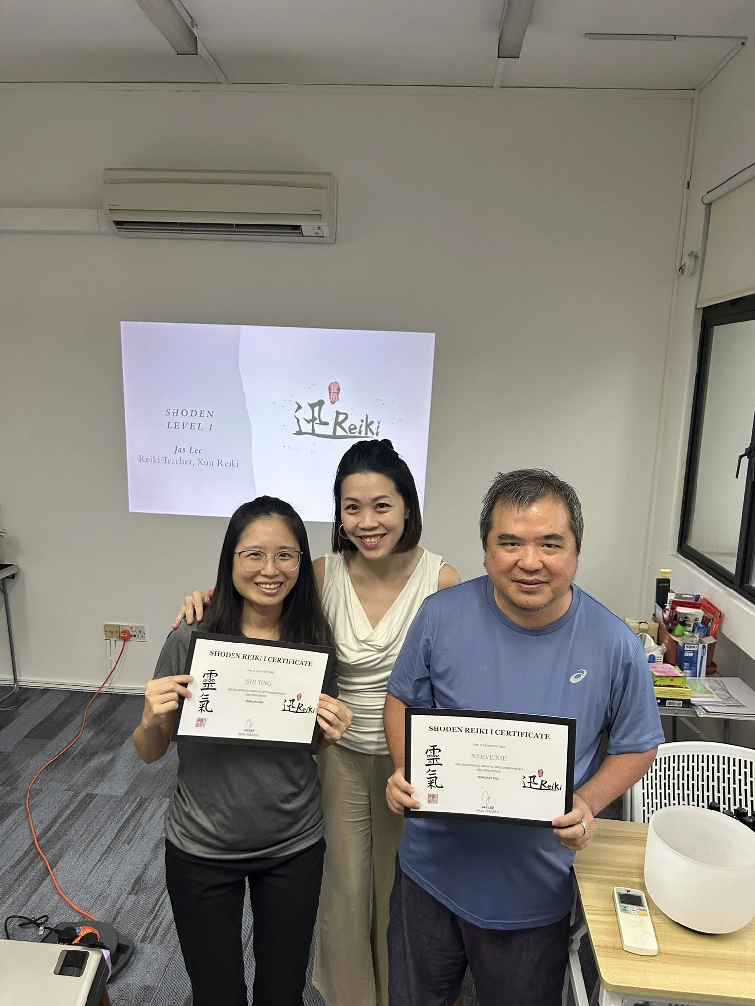 Two women and one man holding certificates in a classroom, with a woman in the middle smiling and two others smiling and standing beside her. A presentation slide about Reiki is visible in the background.