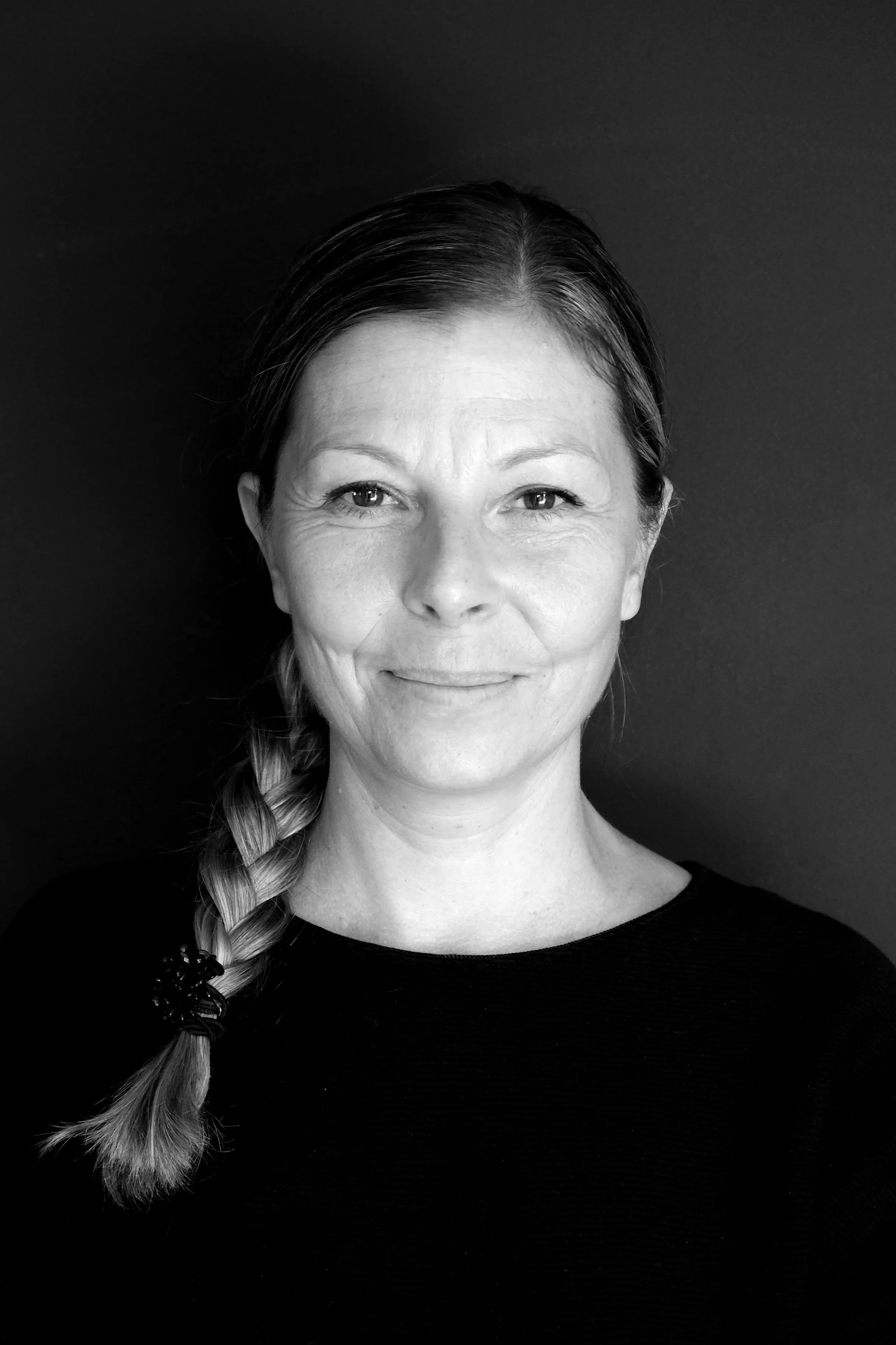 Ritratto in bianco e nero di una donna con capelli raccolti in una treccia, sorriso lieve, sfondo scuro.