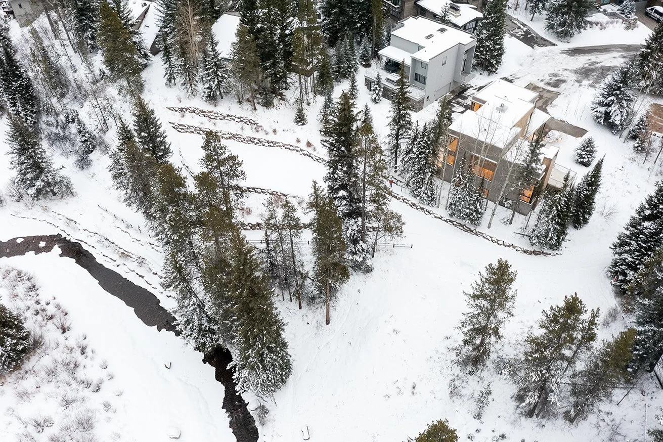 Aerial view of a snowy suburban neighborhood with houses, tall trees, a winding creek, and snow-covered lawns.
