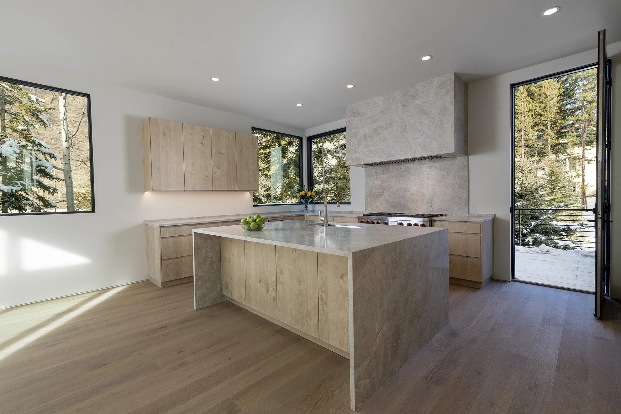Modern kitchen with light wood cabinets, marble island, and large windows showing snowy trees outside.