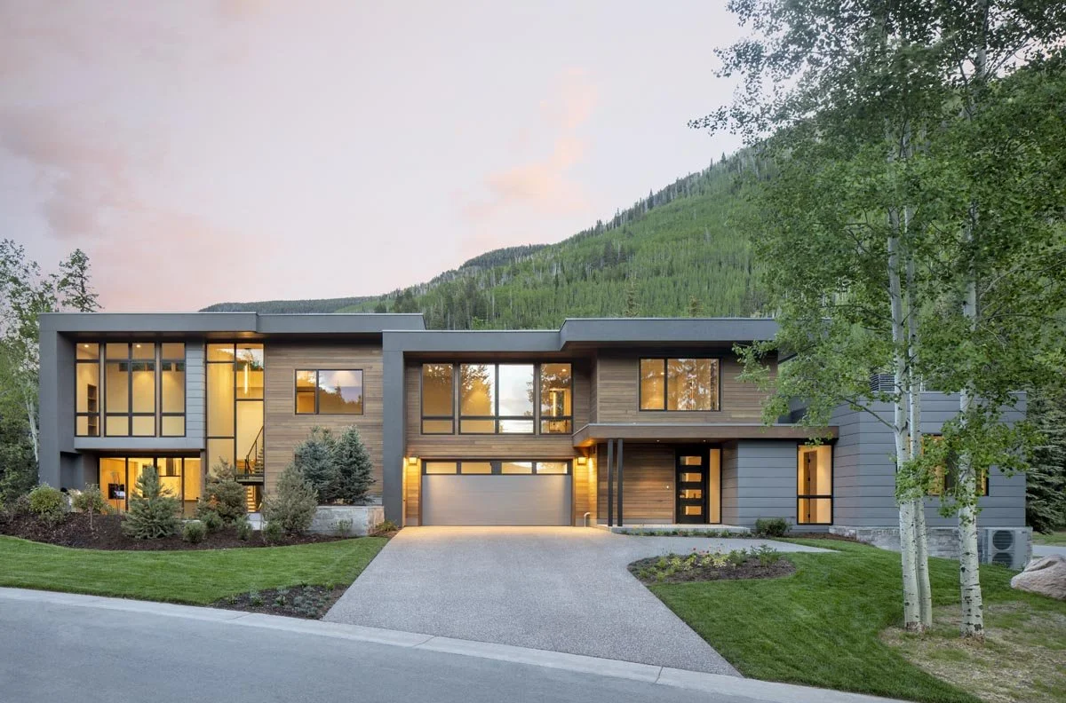 Modern two-story house with large windows, surrounded by trees, with a green lawn and mountain in the background at sunset.