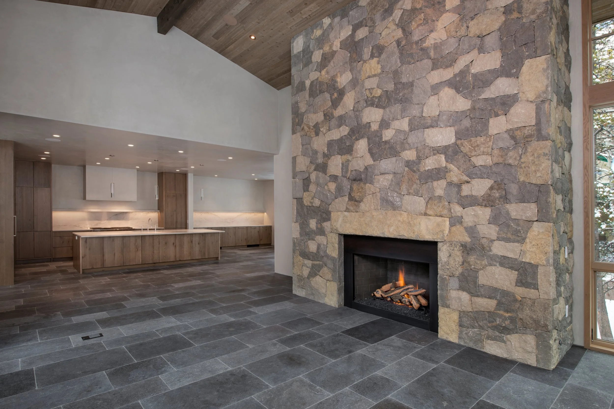 Modern living room with a stone fireplace, gray tile floors, wood-paneled ceiling, and a kitchen area with wooden cabinets and white countertops in the background.