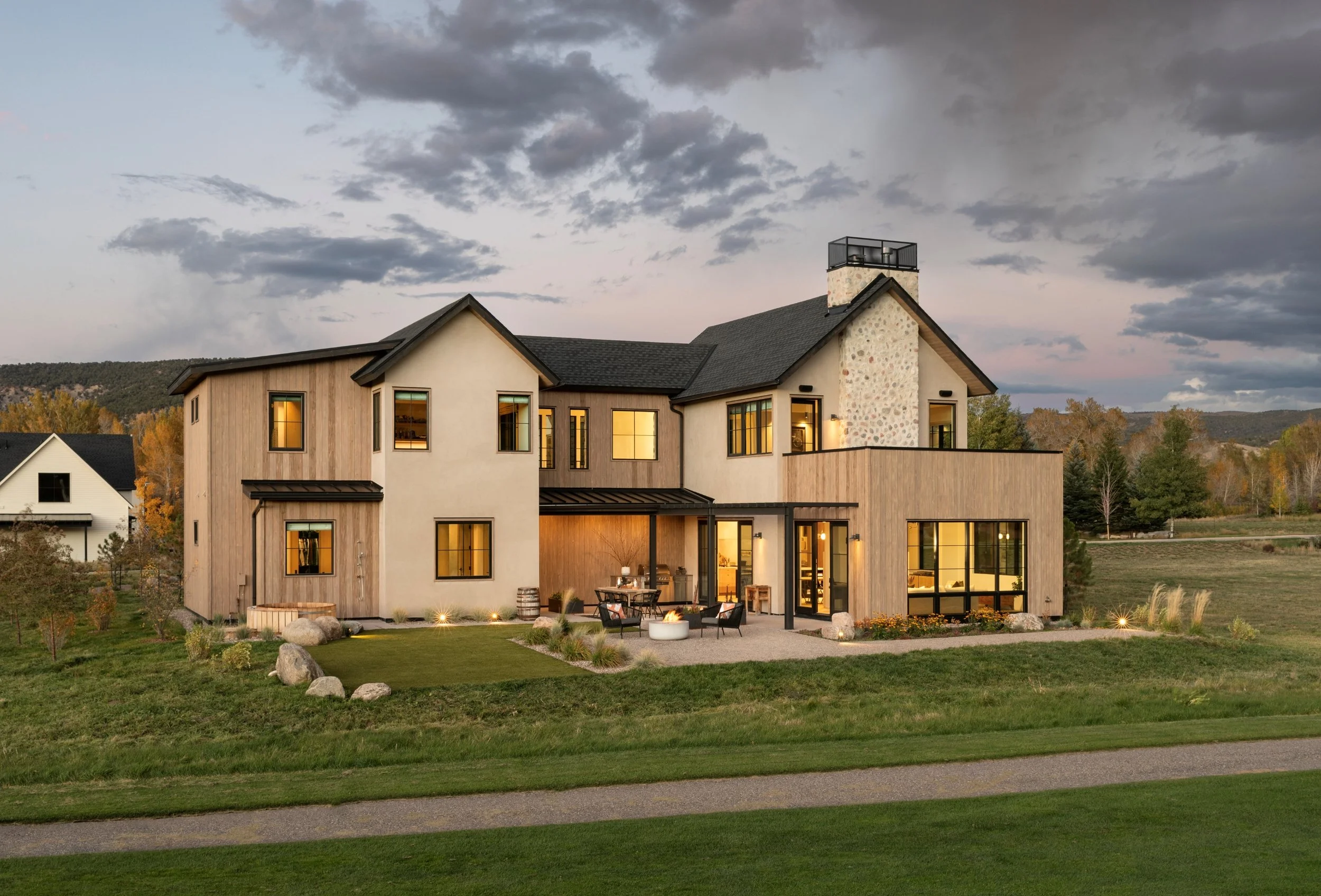 Modern two-story house with large windows, surrounded by a landscaped lawn and a patio with outdoor furniture, under a cloudy evening sky.