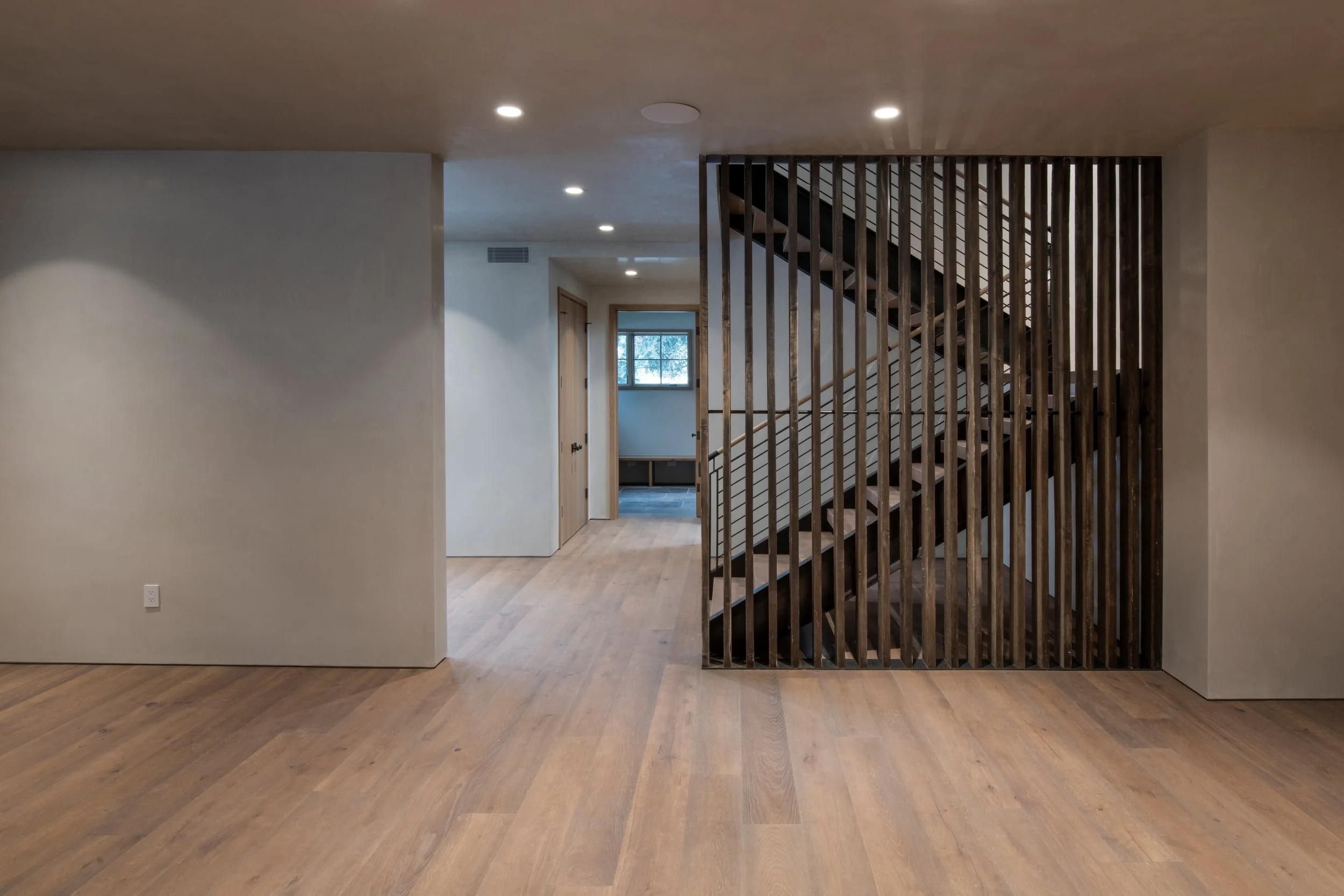 Interior view of a modern home with wooden floors, a staircase enclosed in vertical wooden slats, and a hallway with doors leading to other rooms.