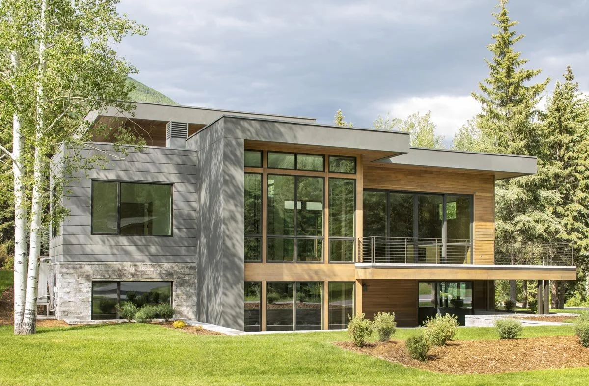 Modern house with large windows, gray and wood exterior, surrounded by green grass and trees.