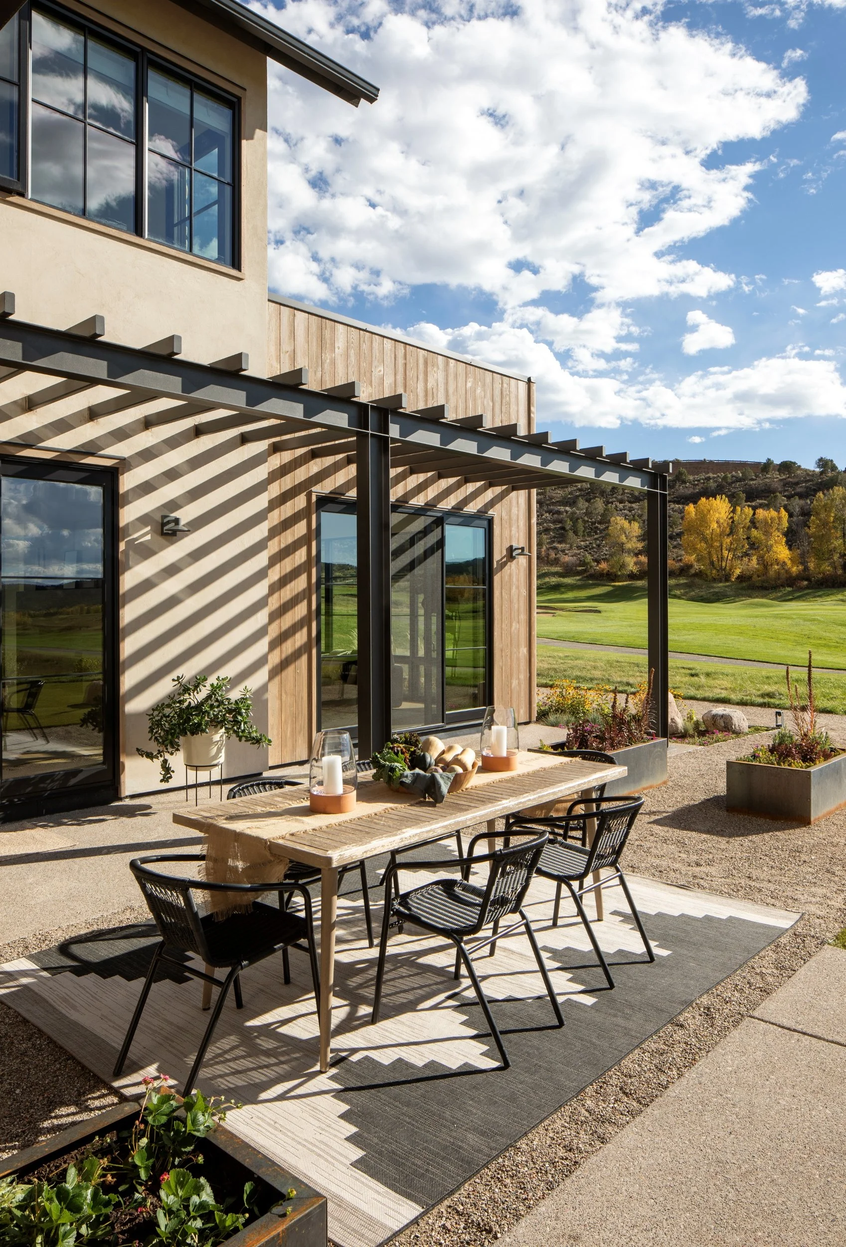 Outdoor dining area with a wooden table, black chairs, and decorative items on a patio with a striped shade pattern. Background features a green landscape and a blue sky with scattered clouds.