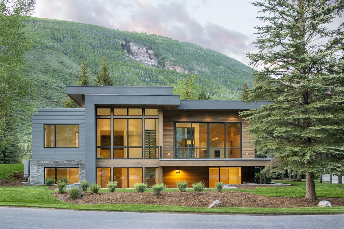 Modern house with large windows, surrounded by greenery and trees, with a mountain backdrop during dusk.