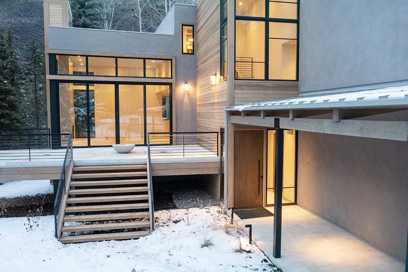 Modern house with large glass windows, wooden accents, and a snow-covered yard, illuminated by exterior lighting at dusk.