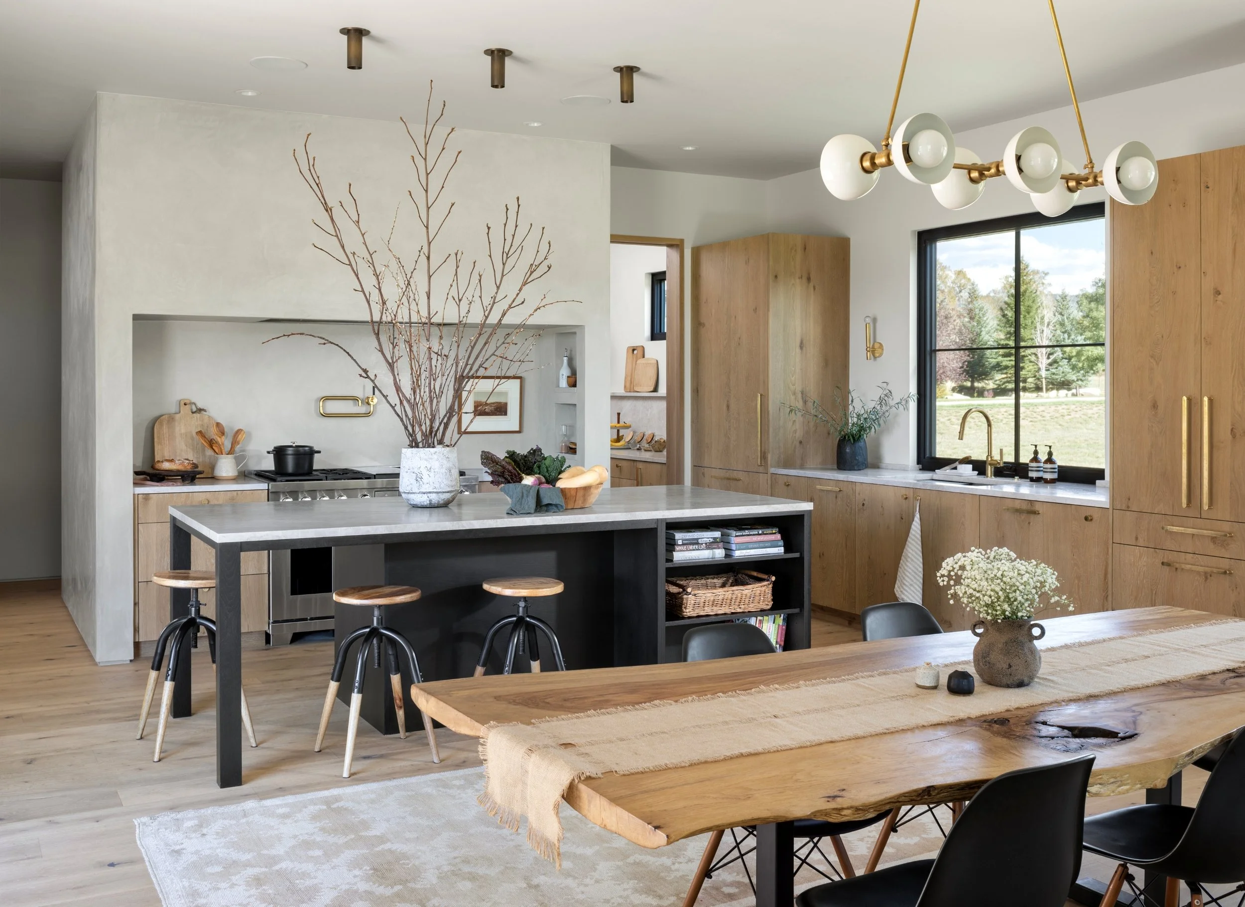 Modern kitchen with wooden cabinets, a kitchen island with a gray base, a large wooden dining table with a beige table runner, black chairs, and decorative items including flowers and pottery. Large window with a rural view, and a contemporary chande