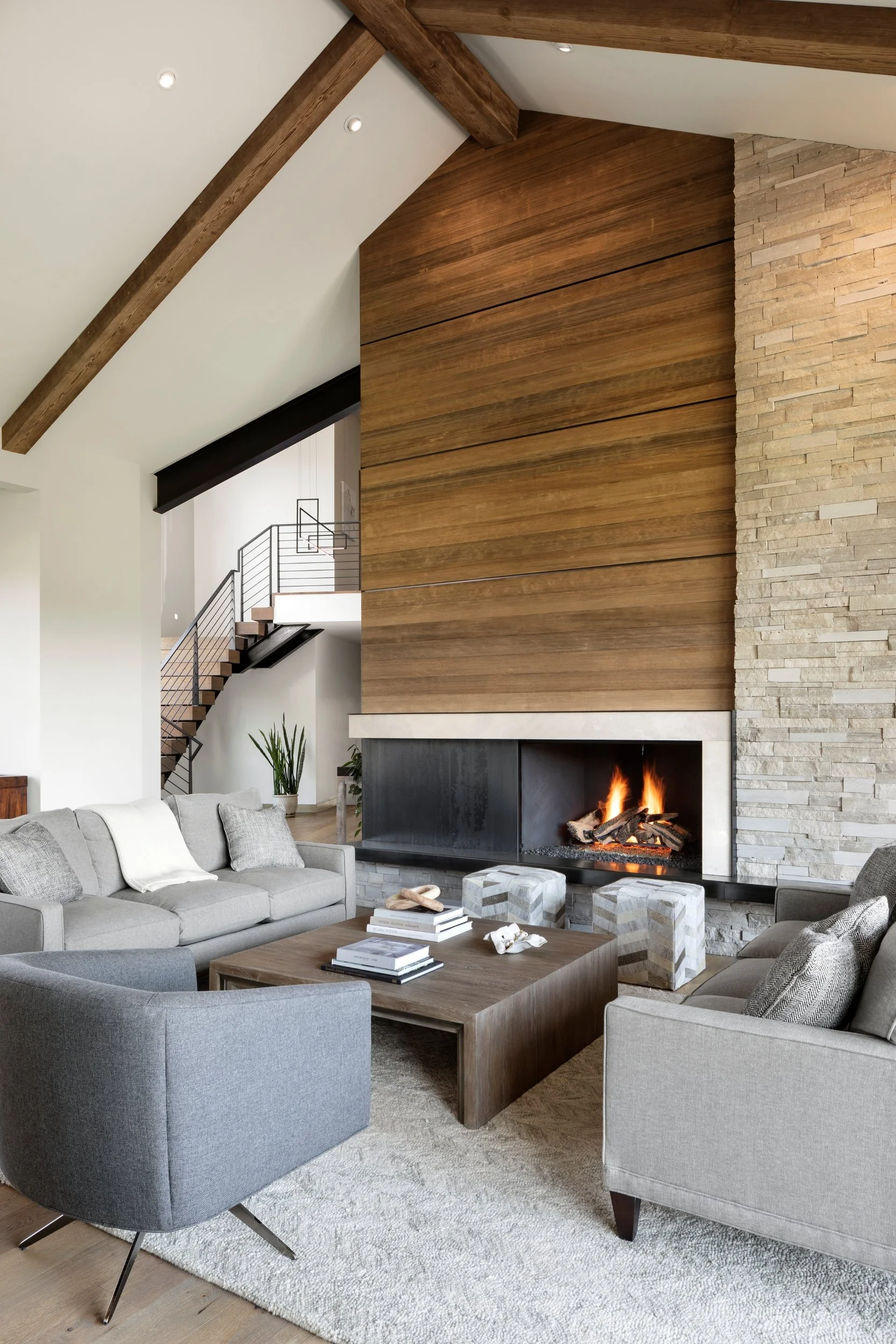 Modern living room with a wood-paneled wall, a fireplace, gray couches, a wooden coffee table, and a staircase with a metal railing in the background.