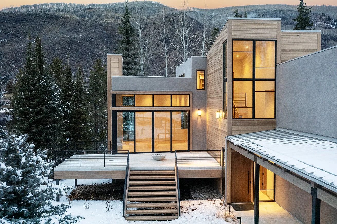Modern house with large windows in a snowy, mountainous landscape at dusk.