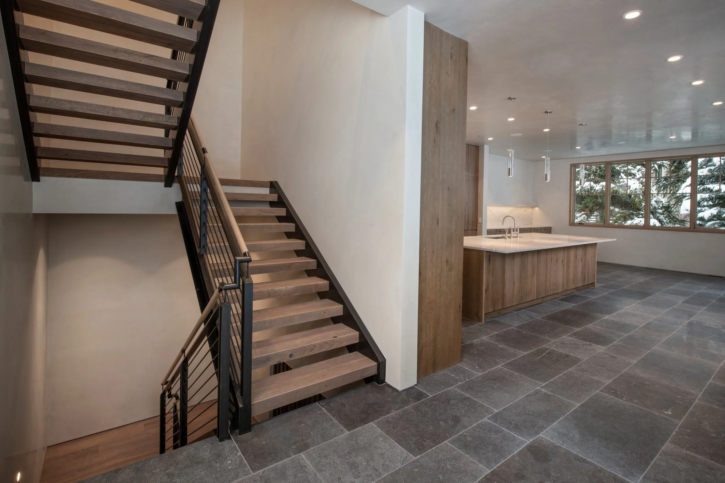 Modern open-concept kitchen with wooden cabinets, a large island, and a window overlooking trees. A staircase with black metal railing and wooden steps leads to an upper level. Recessed lighting and pendant lights illuminate the space, which has a ti