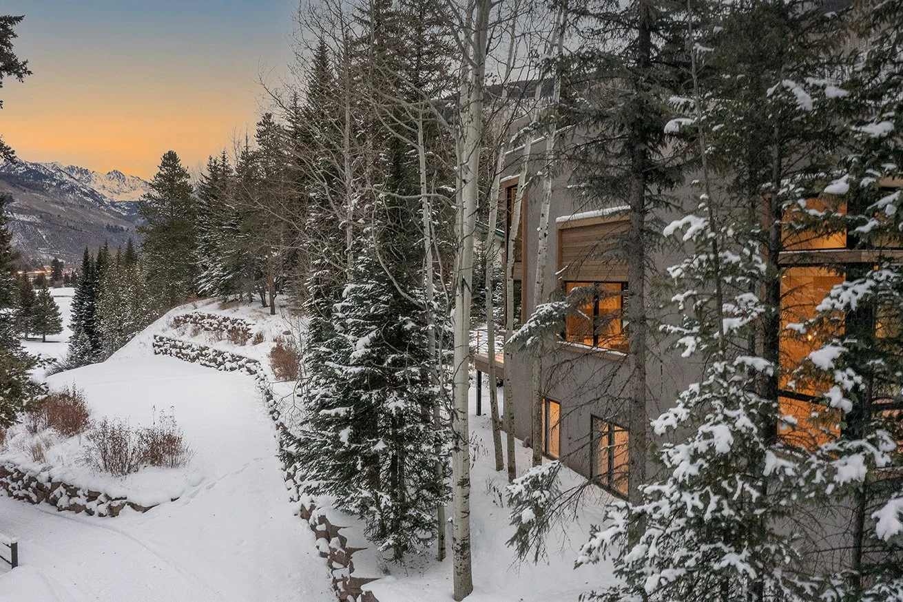 Snow-covered landscape near a multi-story building with lit windows, surrounded by pine trees and mountain in the background during sunset.
