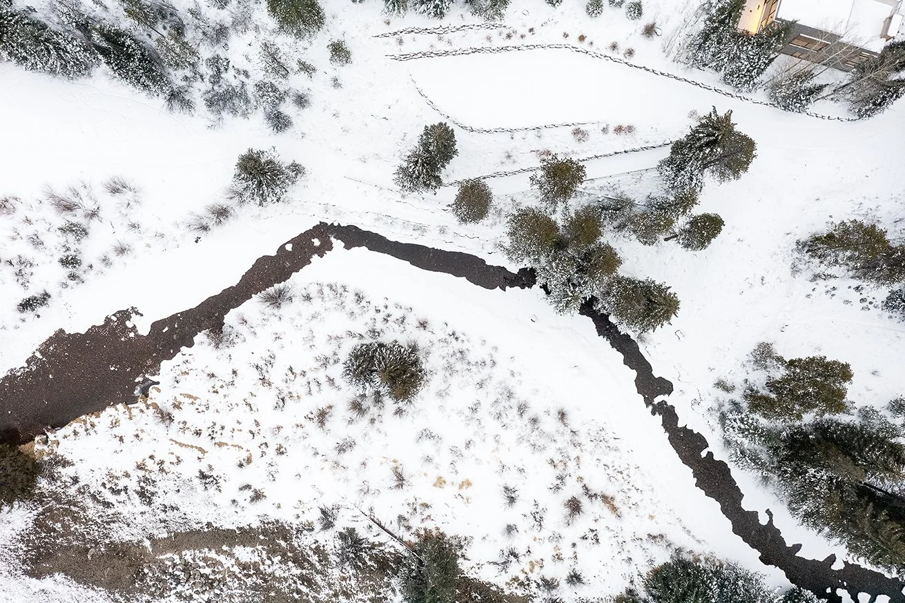 Aerial view of a snow-covered landscape with a winding, narrow creek and scattered trees.