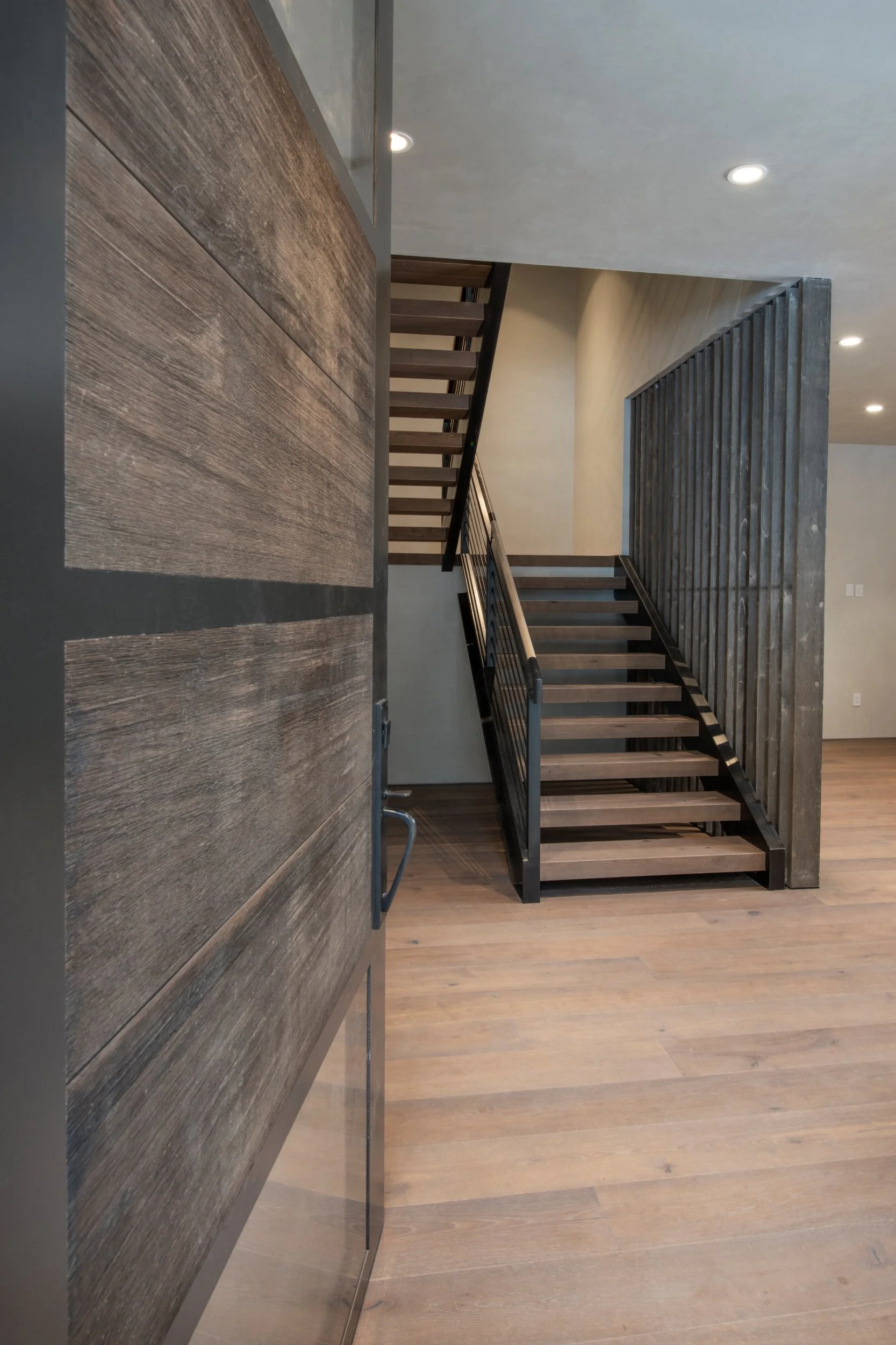 Interior view of a modern staircase with wooden steps and metal railings, adjacent to a textured wooden wall and a vertical wooden slat divider, with a hardwood floor and recessed ceiling lights.