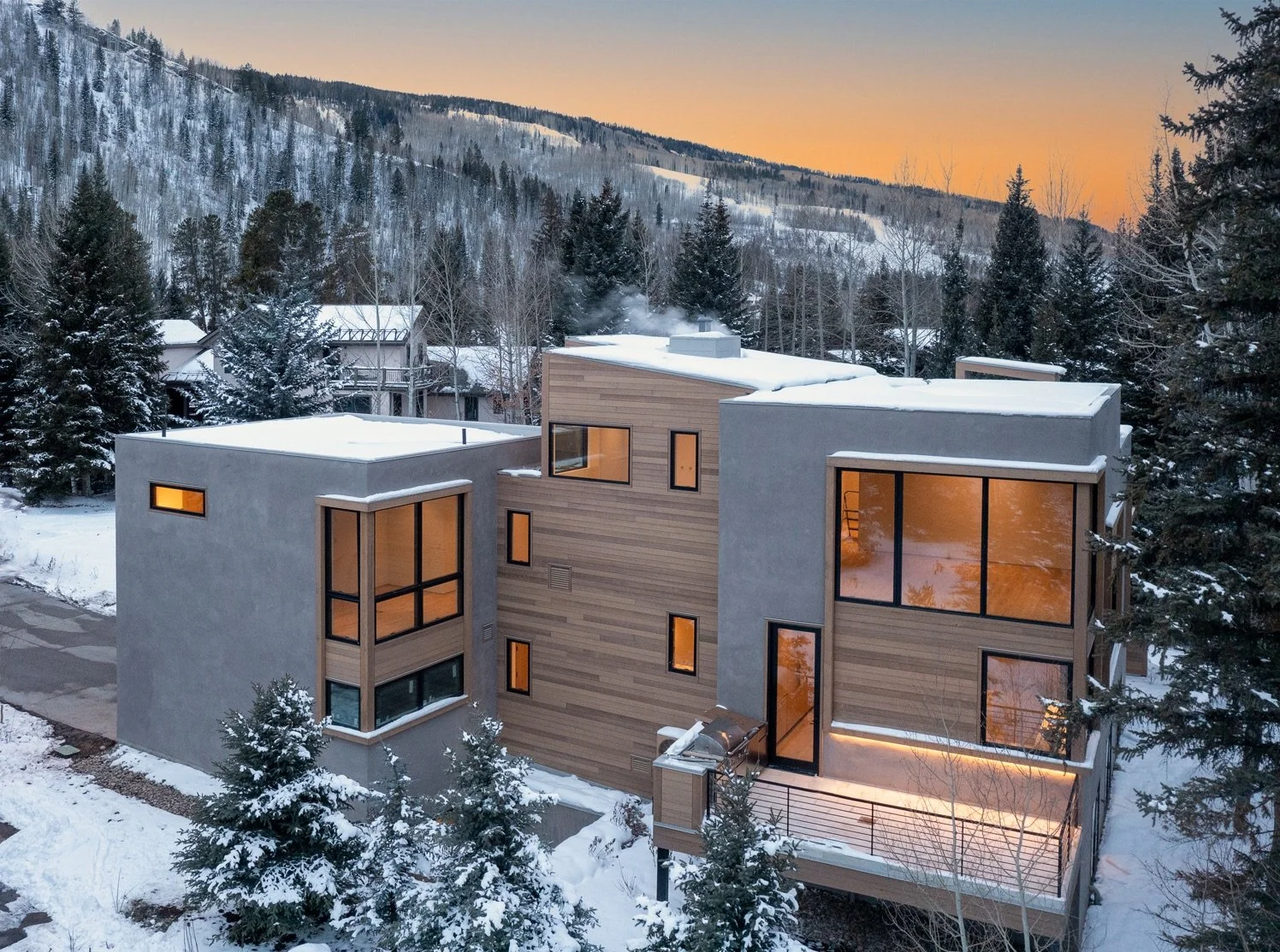 Modern house with large windows, situated in snowy landscape with trees, mountains, and sunset sky in background.