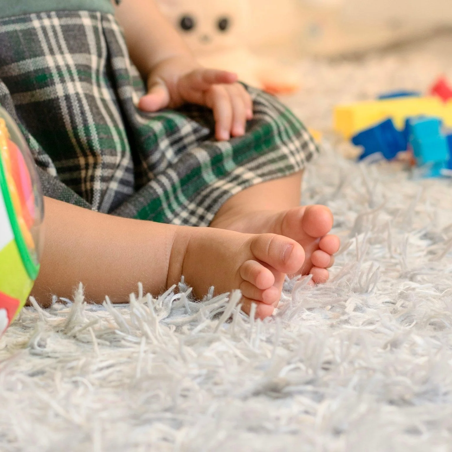 Close-up of a baby's feet resting on a white shaggy rug, with a child's hand nearby and colorful toy blocks in the background.