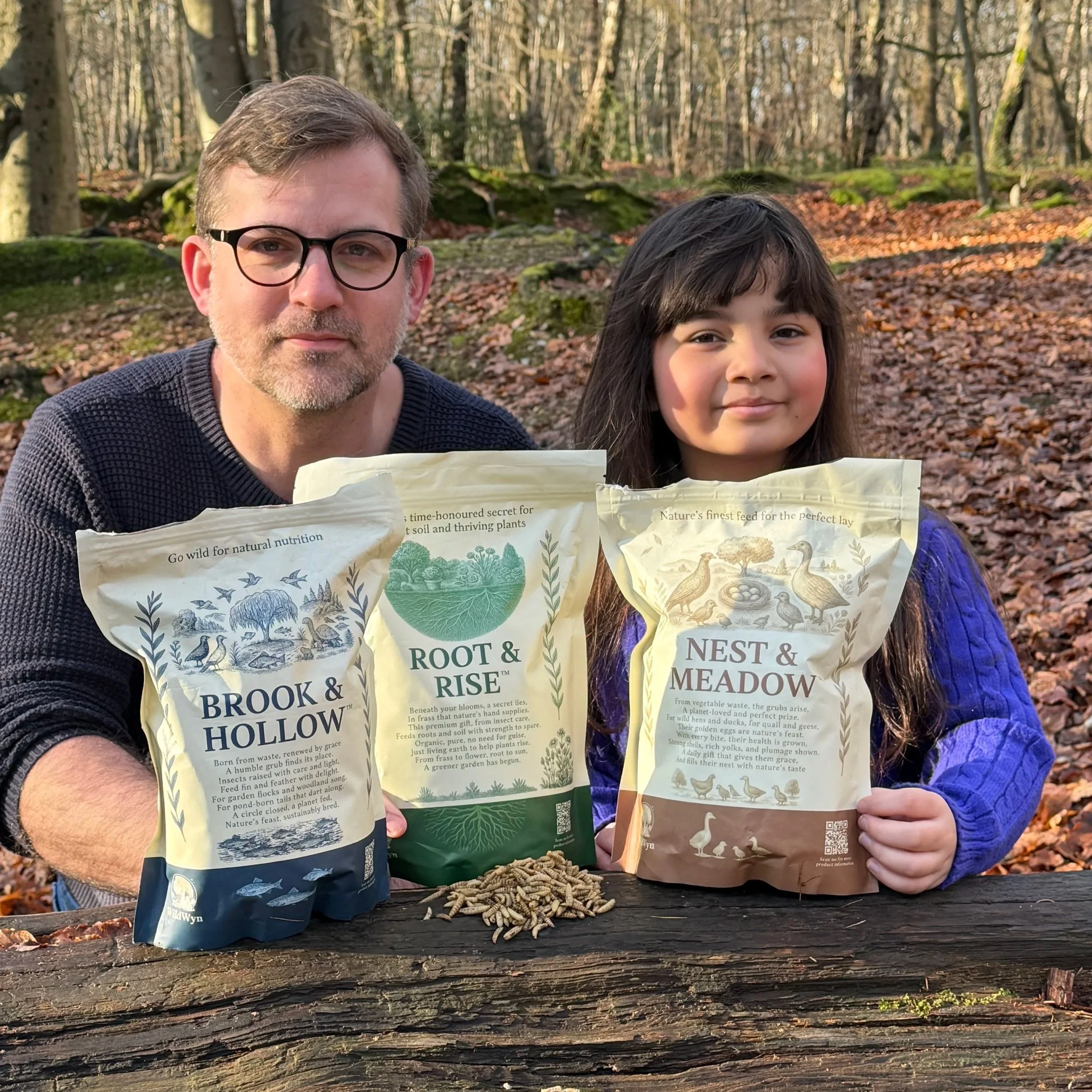 Two people, an adult man and a young girl, holding plant seed packets outdoors in a forest setting with fallen leaves on the ground. The man has glasses and light brown hair, the girl has dark hair with bangs.