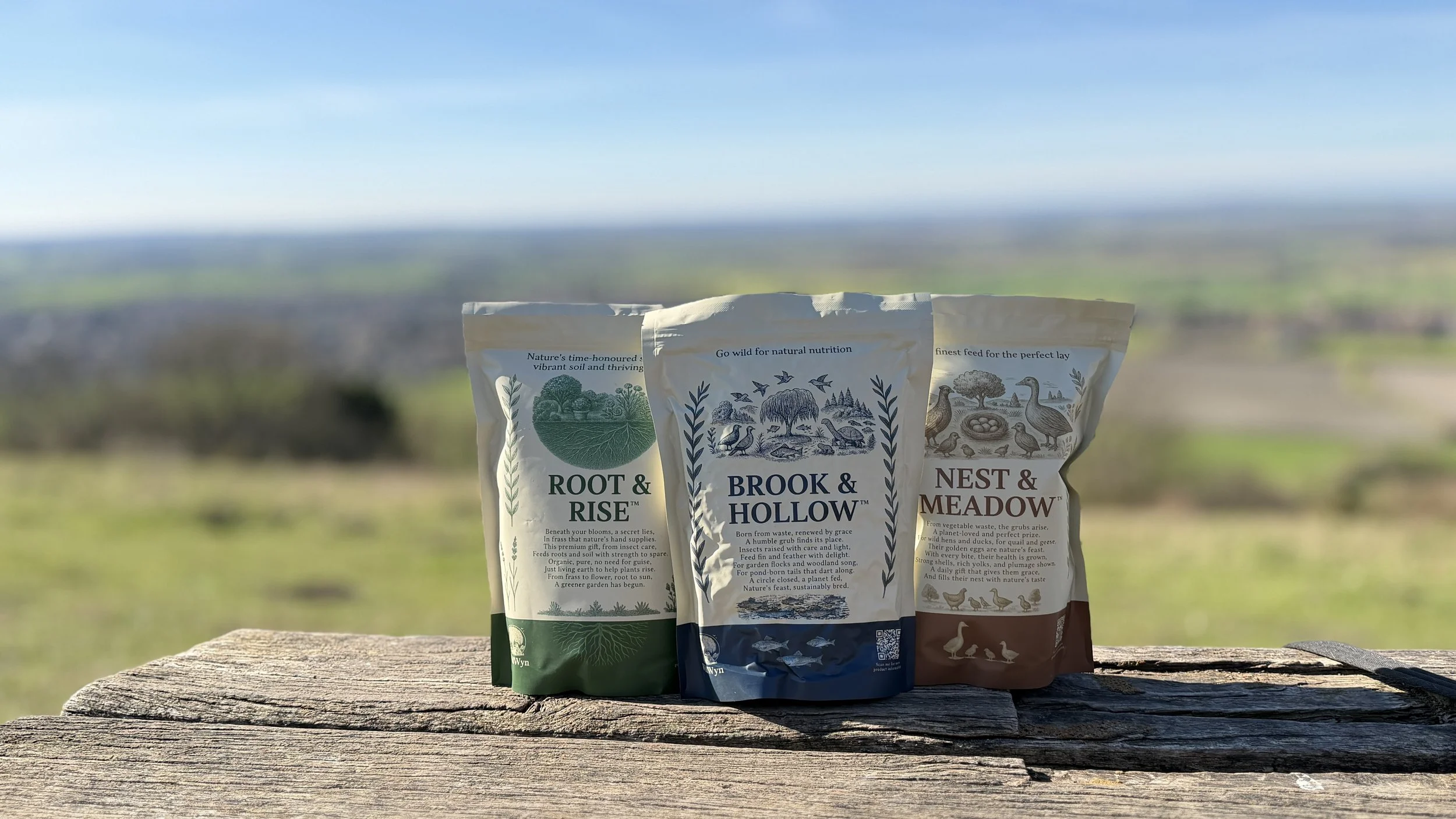 Three bags black soldier fly larvae products 'Root & Rise,' 'Brook & Hollow,' and 'Nest & Meadow' placed on a wooden surface outdoors with a blurred landscape background.