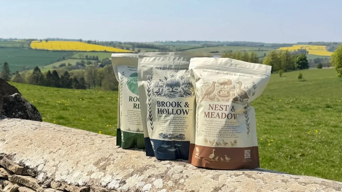 Three bags of bird feed placed on a stone ledge in a rural landscape with green fields, trees, and yellow flowering crops in the distance.