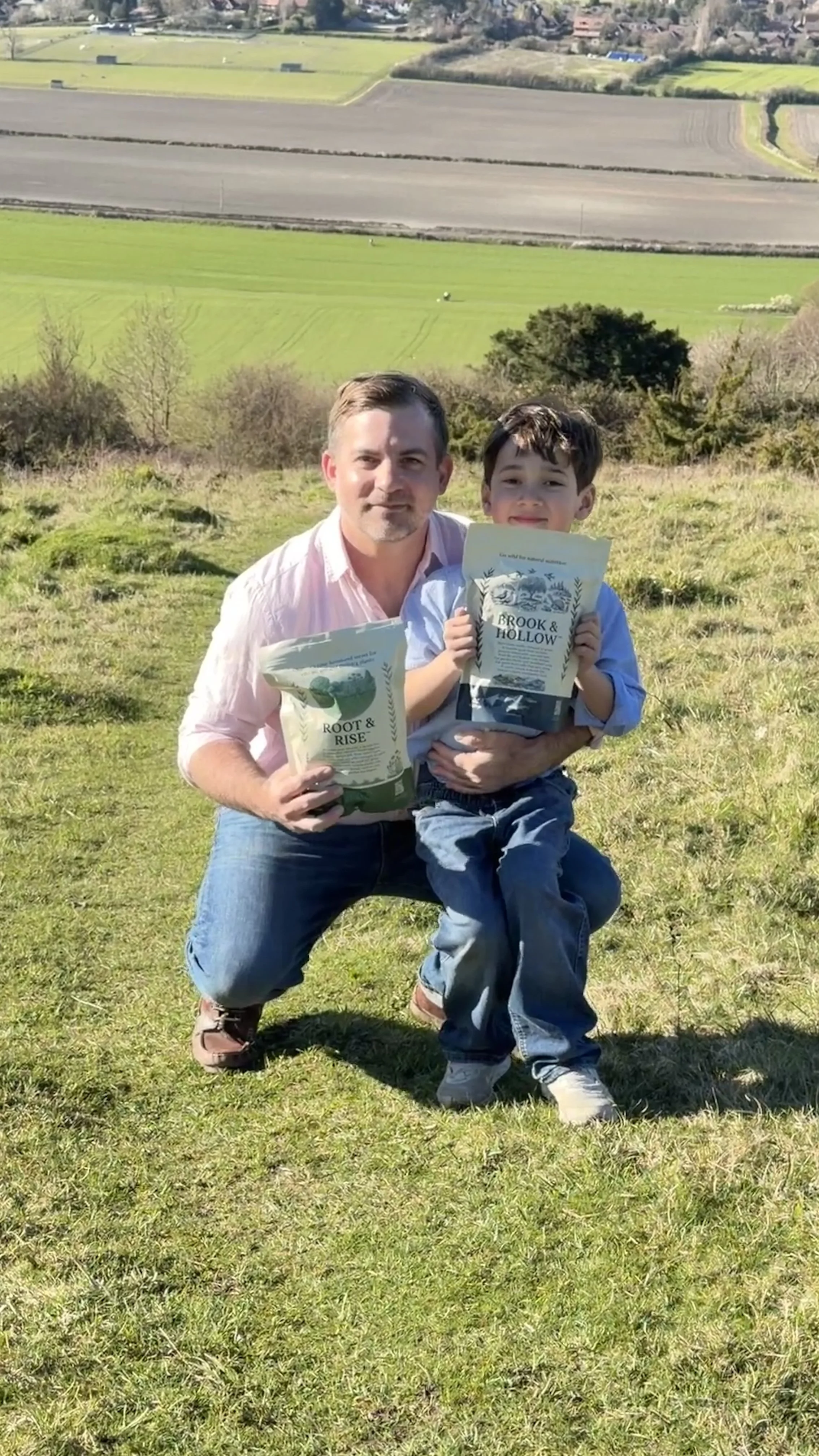 Tom Berry and son are outdoors on a grassy hill, holding books titled "Root & Rise" and "Brook & Hollow" respectively. They are smiling and posing for the photo, with a scenic landscape of fields and trees in the background.
