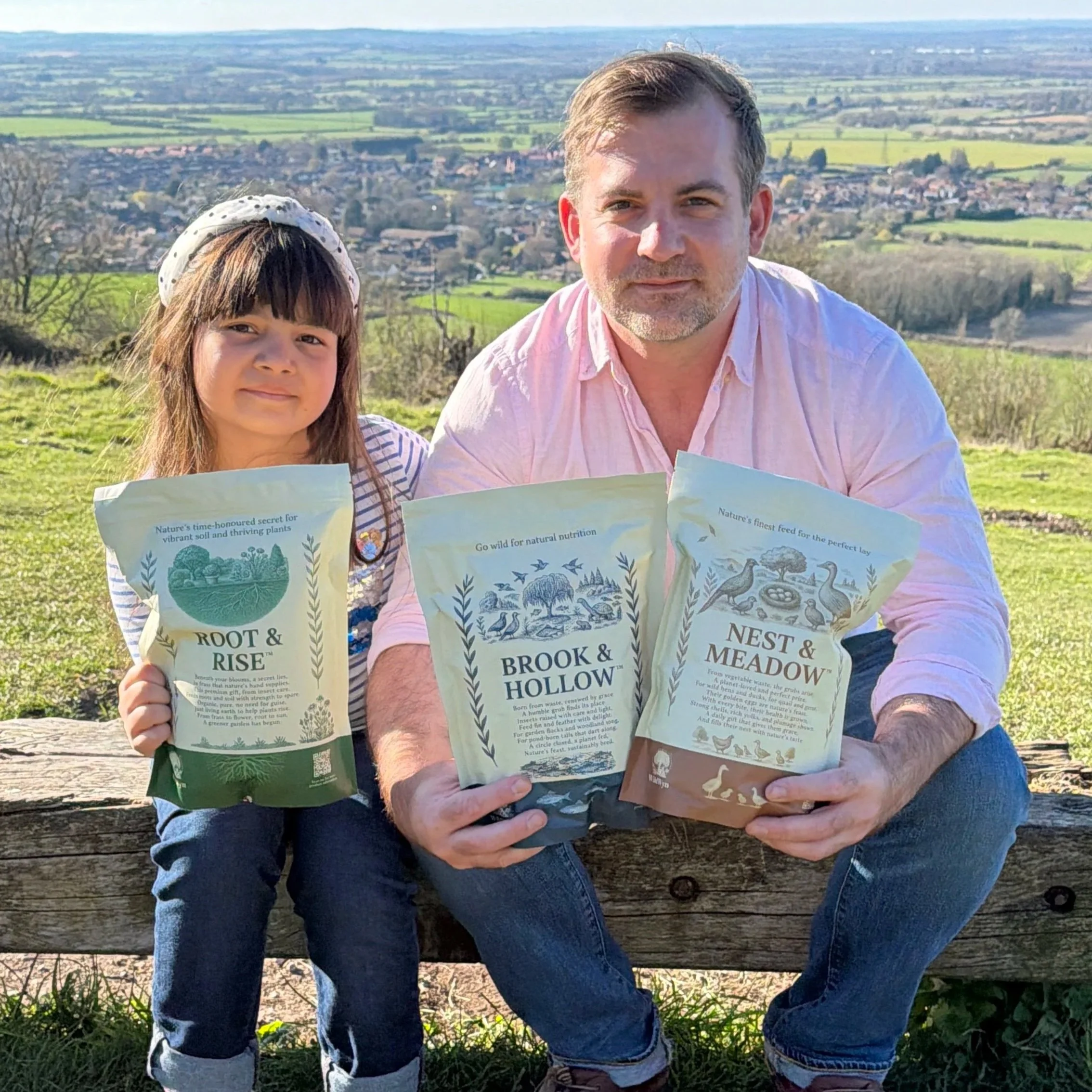 Tom Berry (founder of Wildwyn) sitting outdoors on a wooden bench, holding pamphlets about plants and nature. The background features a vast landscape with fields, trees, and a town, under a clear sky.