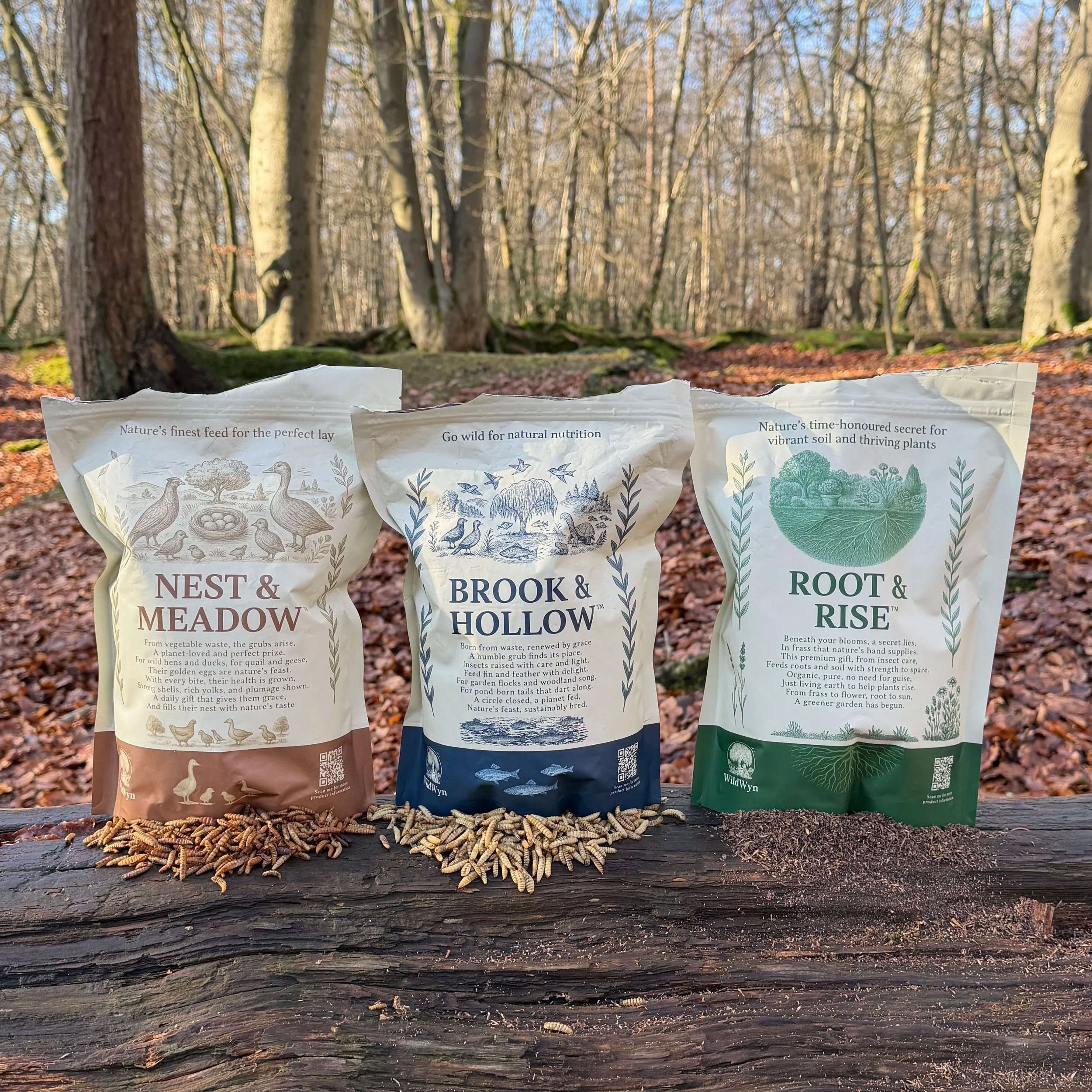 Three packets of insect (black soldier fly larvae products) placed on a fallen tree trunk in a wooded area with trees in the background.