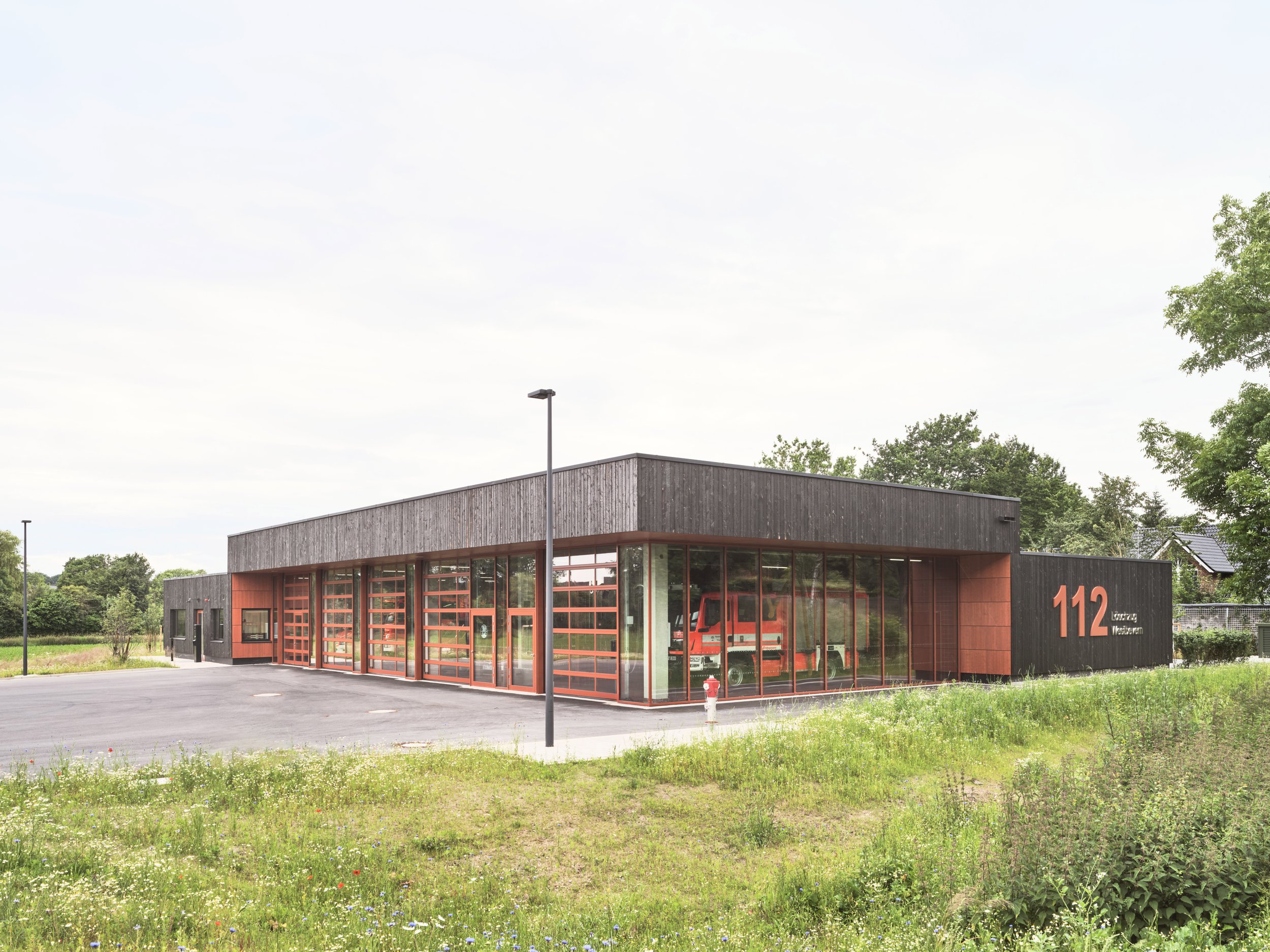 Modernes Feuerwehrgebäude mit großen Glasfenstern und roten oberen Türrahmen, das orange Feuerwehrfahrzeug im Inneren sichtbar, Außenbereich mit Parkplatz und ein paar Bäumen in der Umgebung. Holztafelbau, Holzkonstruktion, karbonisierte Fassade.