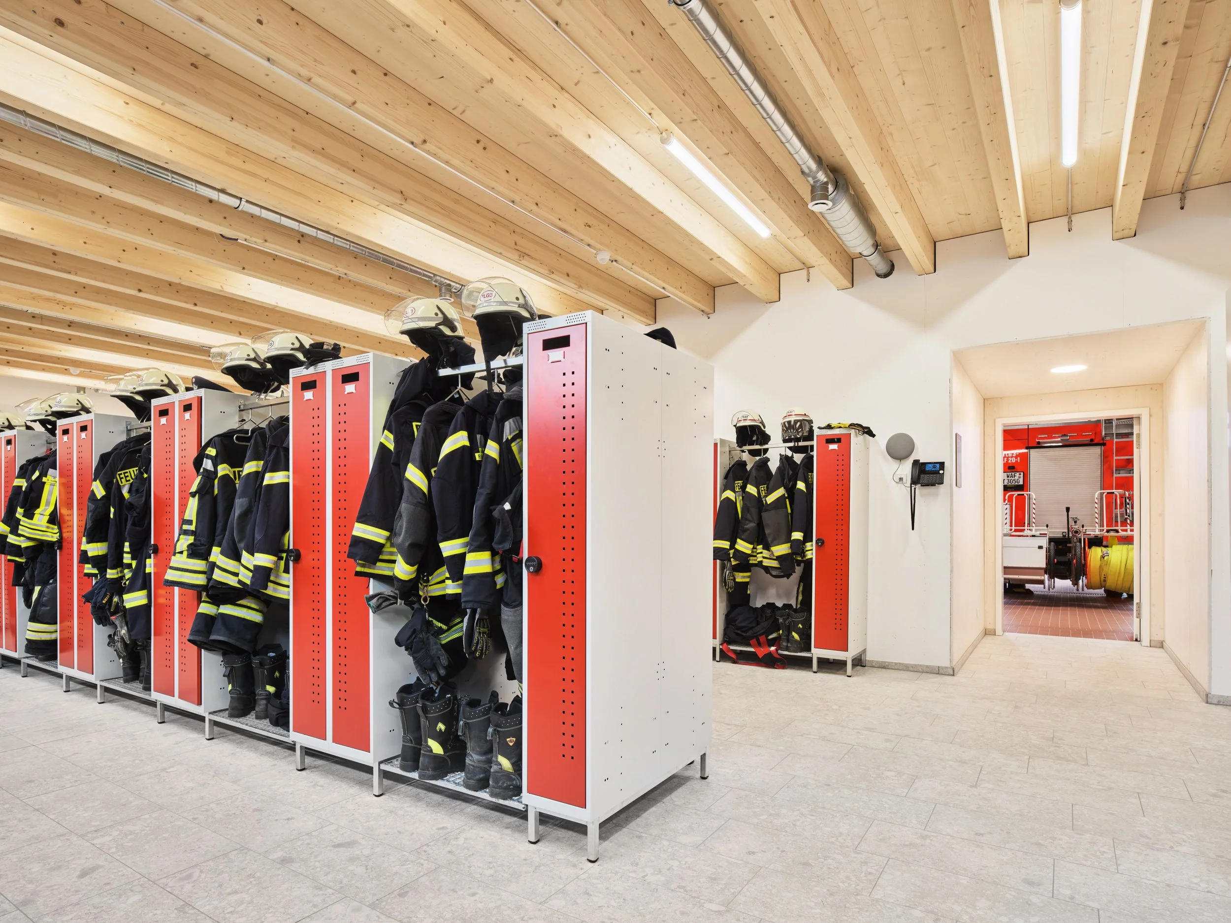 Umkleider der Feuerwache Westbevern. Feuerwehrbekleidung und Helme in Umkleidekabinen, verbunden mit Feuerwehrfahrzeug im Hintergrund.