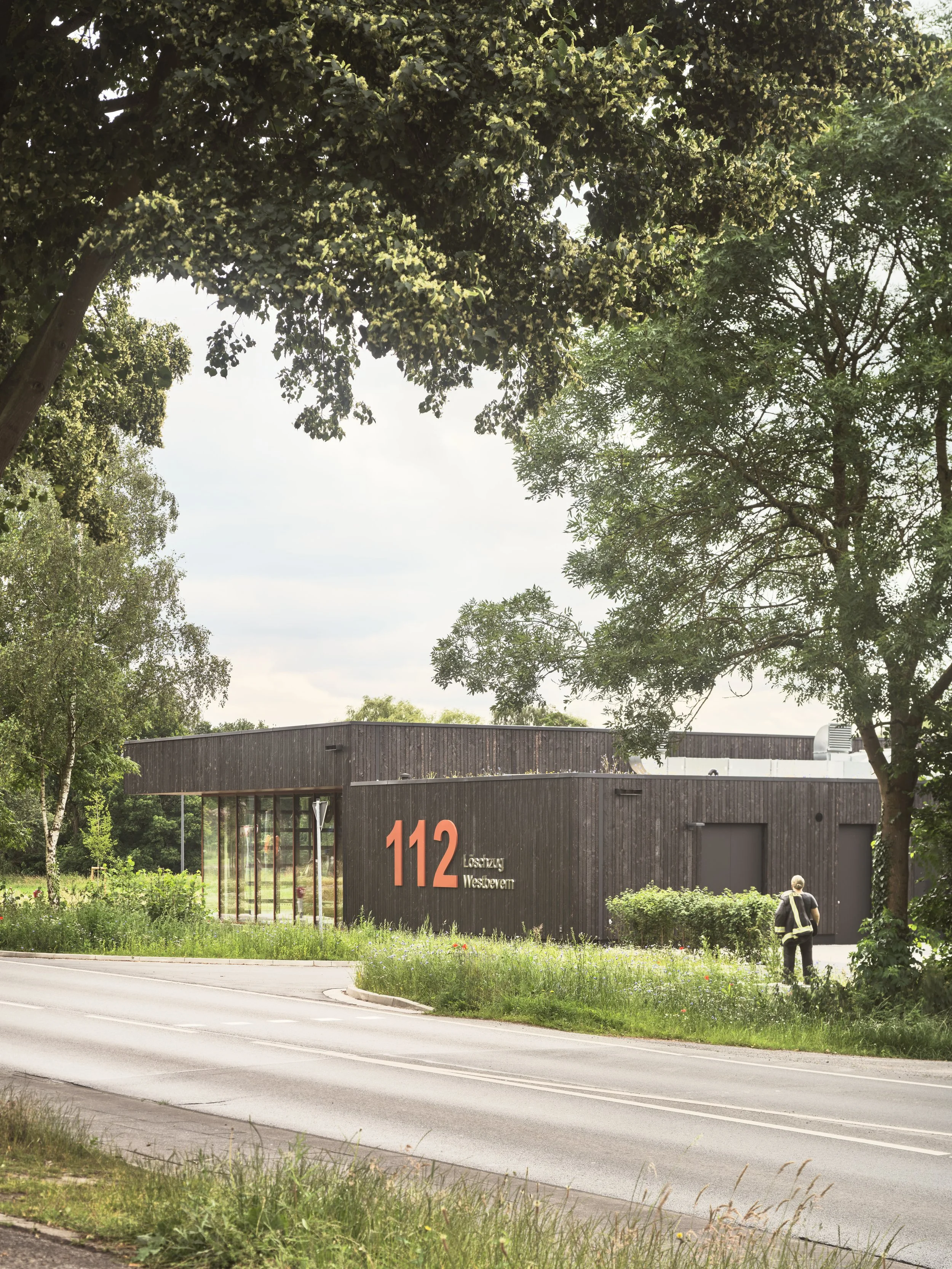 Ein modernes schwarzes Feuerwehr Gebäude mit großen Fenstern und der Nummer 112, umgeben von Bäumen und Gras, an einer Straße, mit einer Person in Arbeitskleidung im Vordergrund. Feuerwache, schwarze Fassade.