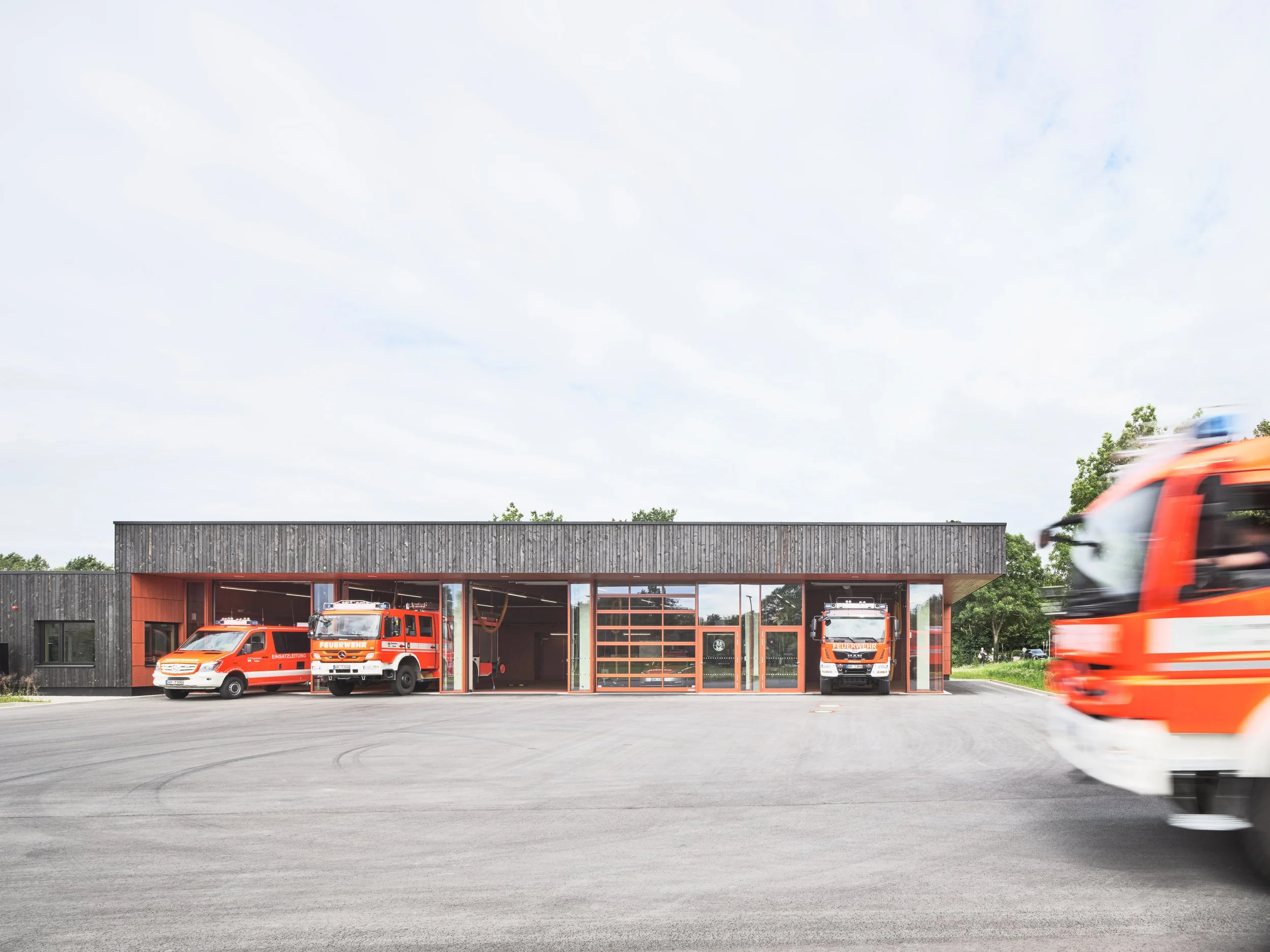 Feuerwache mit drei Einsatzfahrzeugen und einem Feuerwehrauto vor dem Gebäude, im Hintergrund Bäume und Himmel.