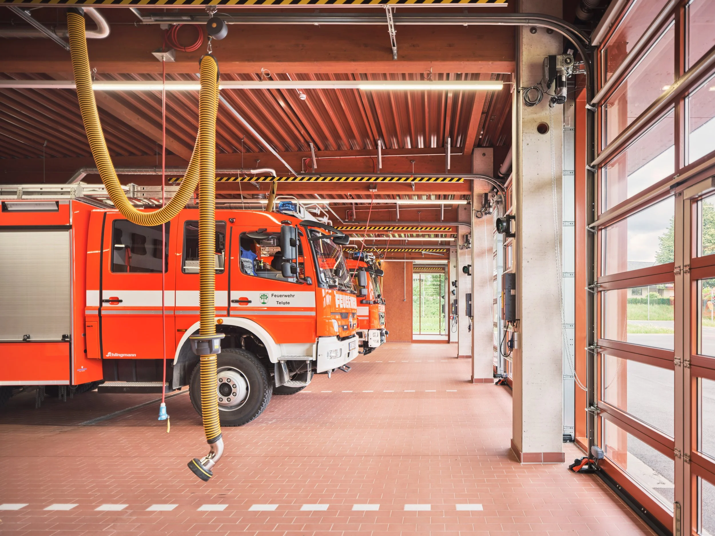 Fahrzeughalle mit rotem Innenraum und großen, verglasten Rolltoren nach draußen. Holztafelbau, Holzkonstruktion, karbonisierte Fassade, Feuerwache.