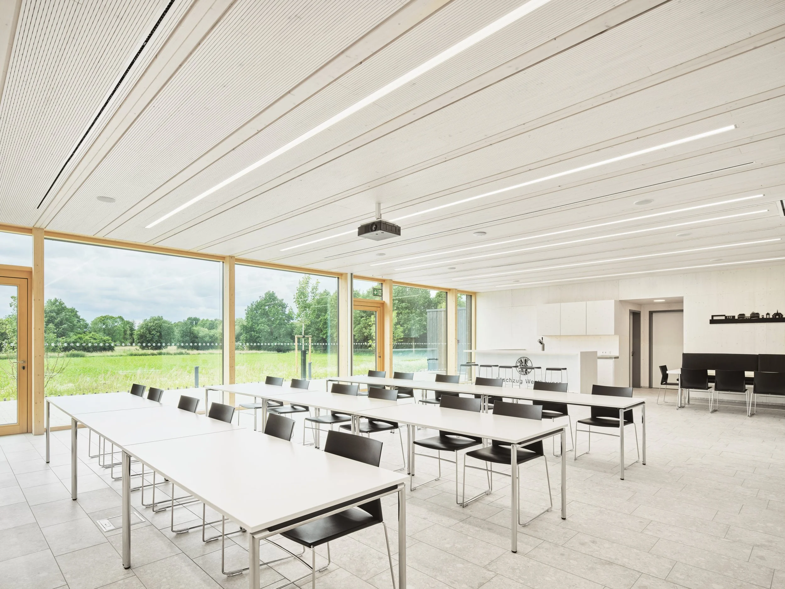 Ein heller, moderner Tagungsraum mit weißen Tischen und schwarzen Stühlen, große Holzfenster mit Blick auf grüne Landschaft, weiße Decke mit integrierten Lichtern, kleiner Küchenbereich im Hintergrund. Holzkonstruktion, karbonisierte Fassade