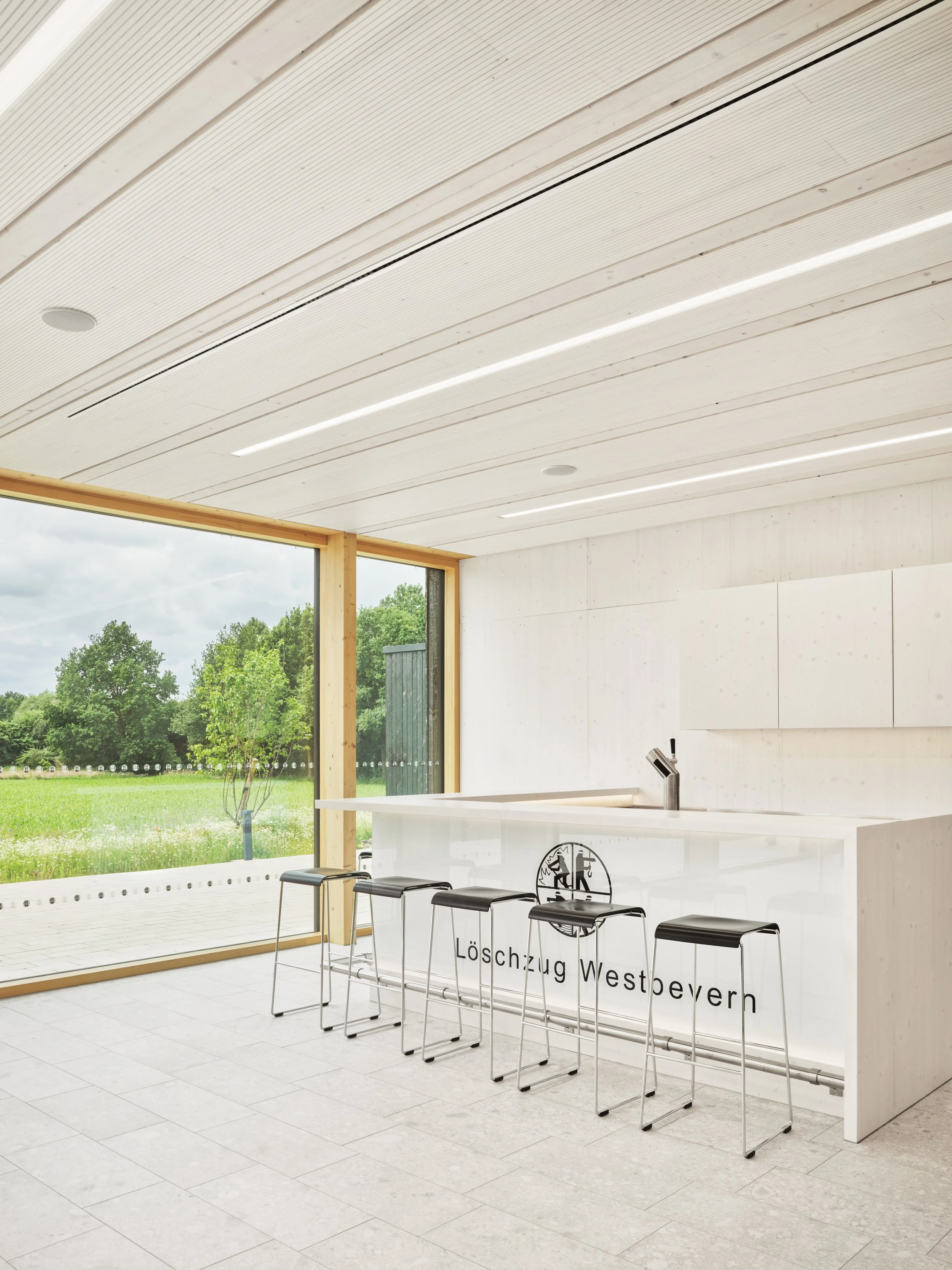 Innenansicht eines modernen, hellen Raums mit großen Fenstern, einem weißen Tresen mit dem Logo 'Löschzug Westbevern' und schwarzen Barhockern davor, Blick nach draußen auf grüne Bäume und Wiese. Holzkonstruktion, karbonisierte Fassade, Feuerwache.