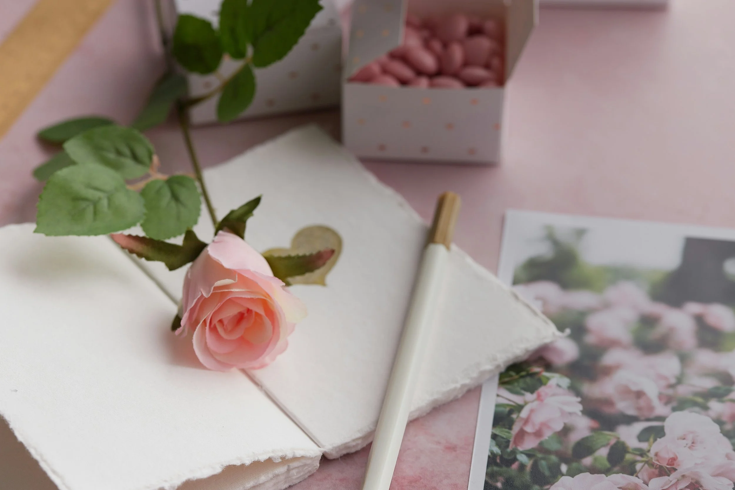 A pink rose with green leaves on a white textured cloth, accompanied by pink chocolates in a patterned white box, a pen, and a photograph of pink flowers, all arranged on a pink surface.