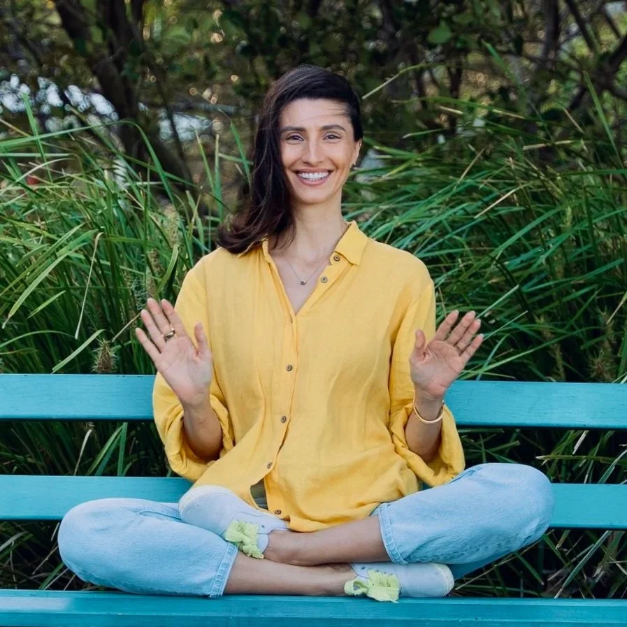 A smiling woman with dark hair sitting cross-legged on a teal bench outdoors, surrounded by green plants. She is wearing a yellow shirt, light jeans, and yellow slippers, with her hands raised in a welcoming gesture.