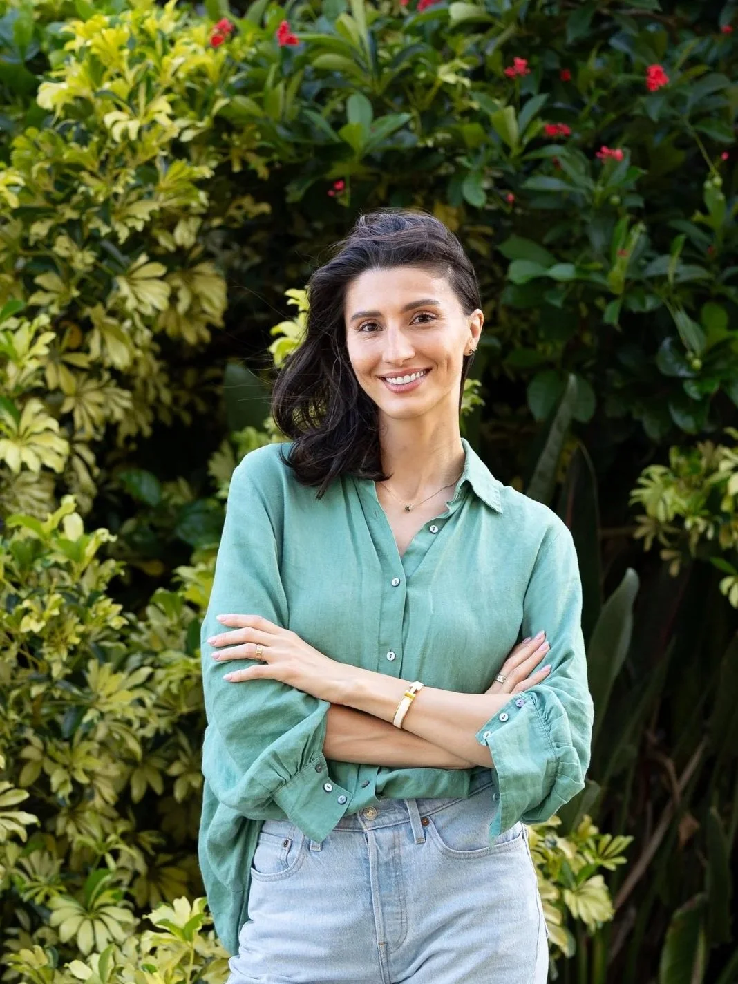 A woman smiling outdoors with her arms crossed, standing in front of green leafy plants and pink flowers.
