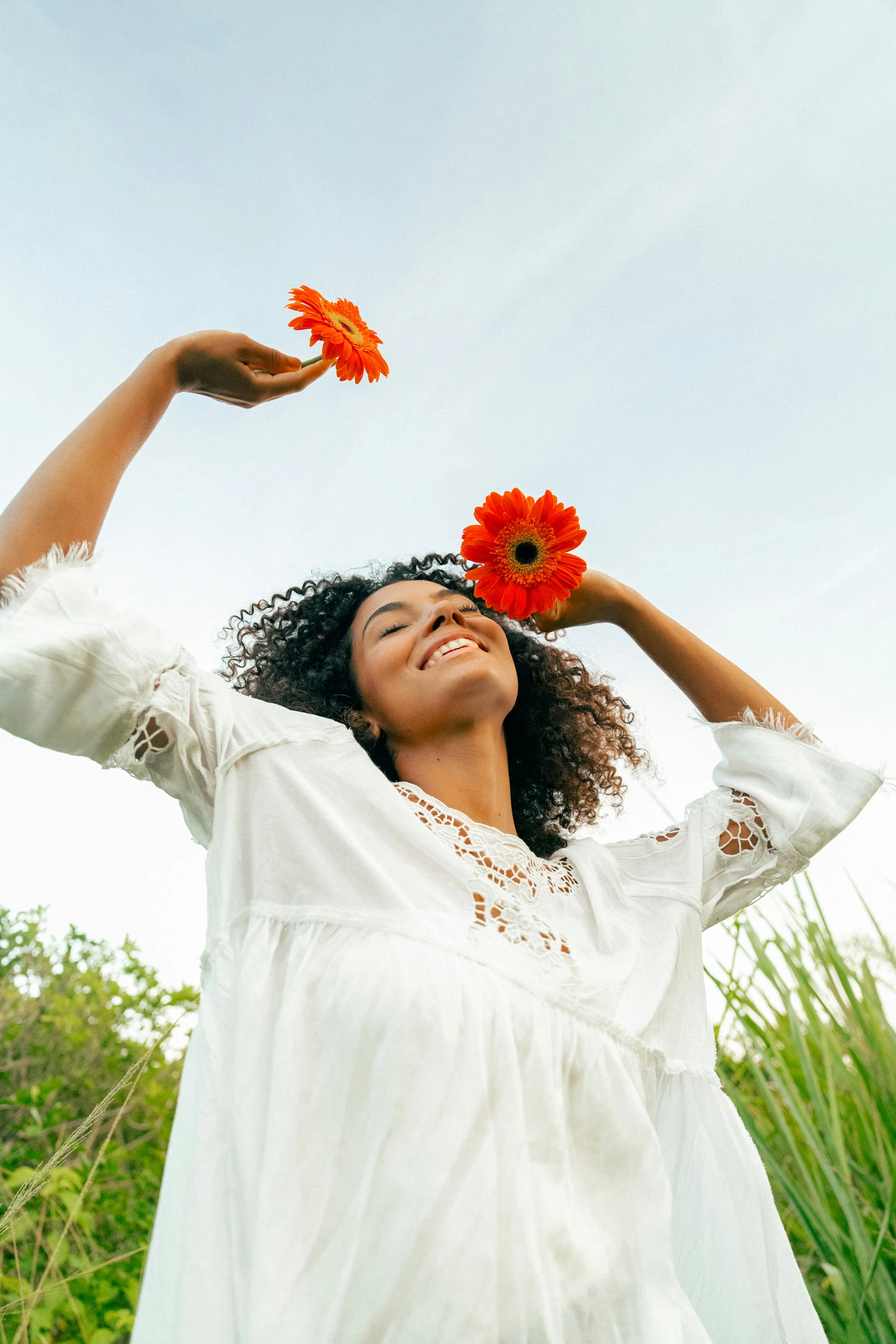 A woman enjoying outdoors holds two orange daisies above her head against a blue sky, smiling with her eyes closed, wearing a white dress with lace details - it emanates freedom, peace and love.