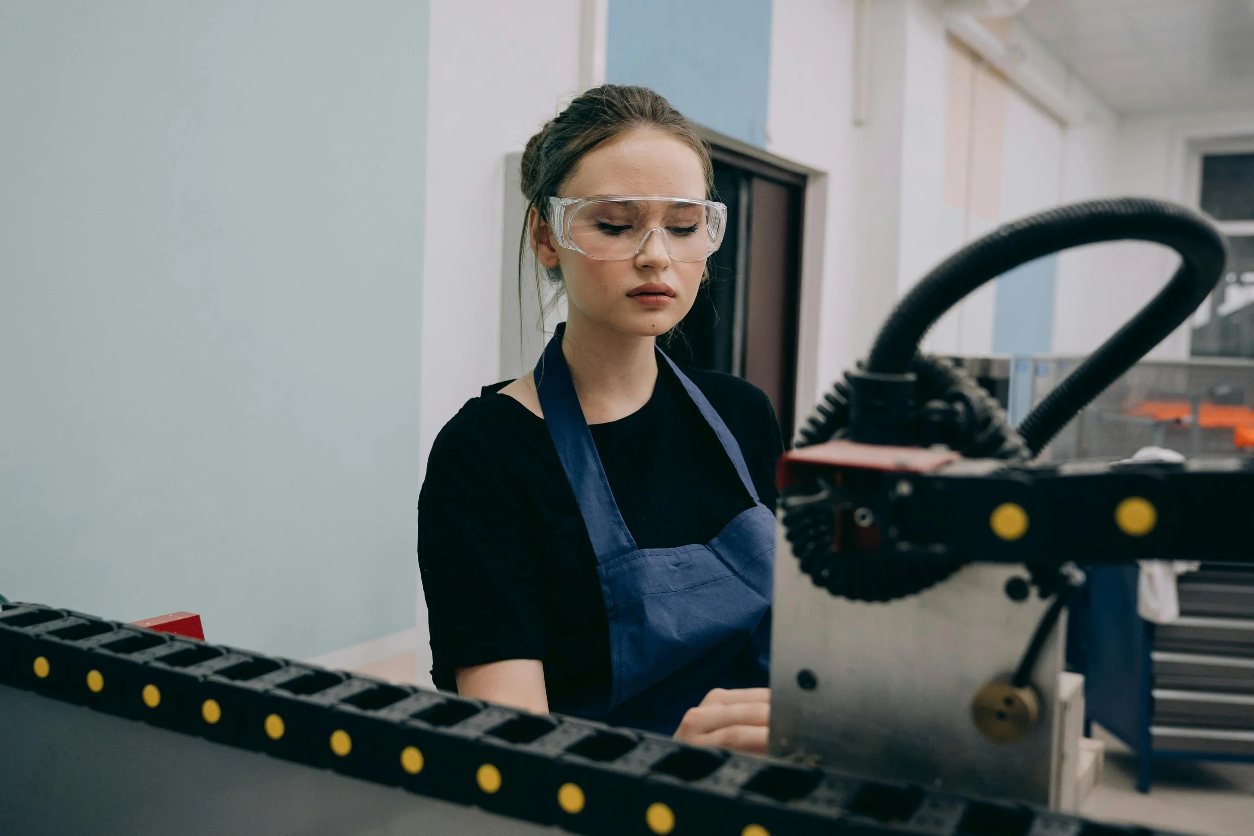 A woman in safety glasses and a black shirt with a blue apron working on a machine with industrial equipment in a factory or workshop setting.