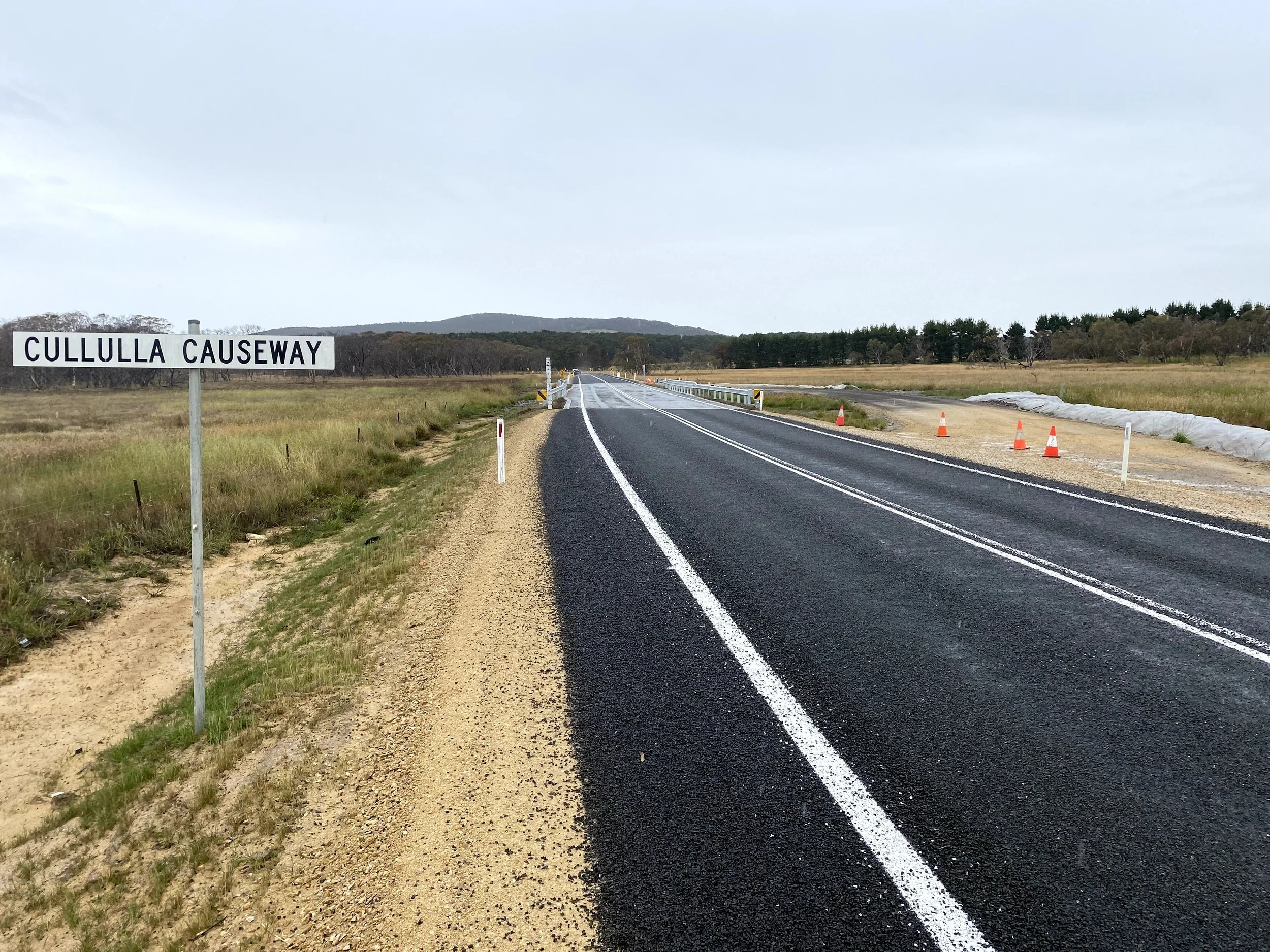A rural road with a sign that reads 'Cullulla Causeway', the road is partly under construction with traffic cones on the side, open fields, trees, and hills in the distance under a cloudy sky.